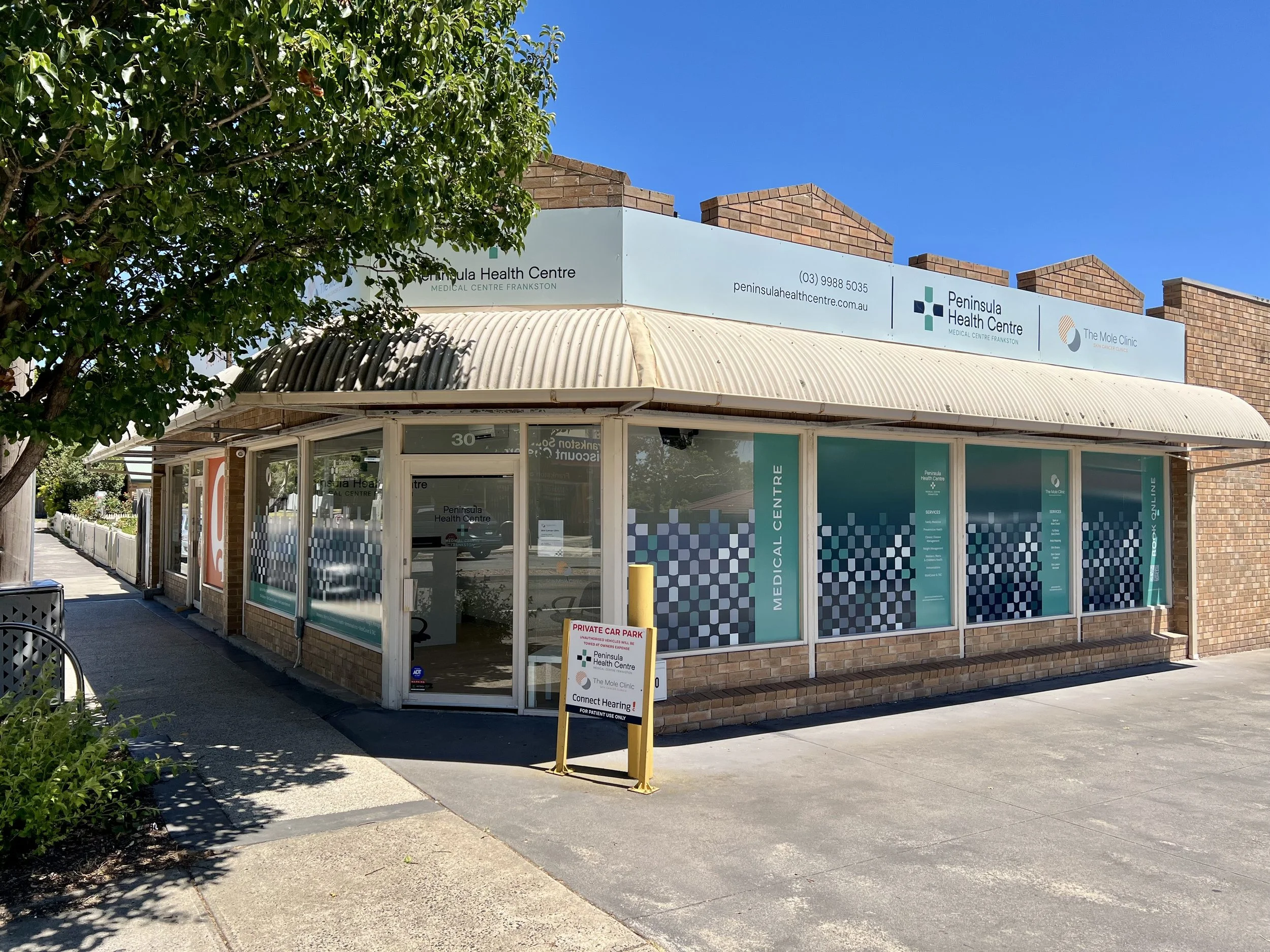 A corner view of Peninsula Health Centre, a medical clinic in a brick building, with blue sky and a tree on the left, and a parking sign in front reading 'Private Car Park' and 'Connect Hearing'.