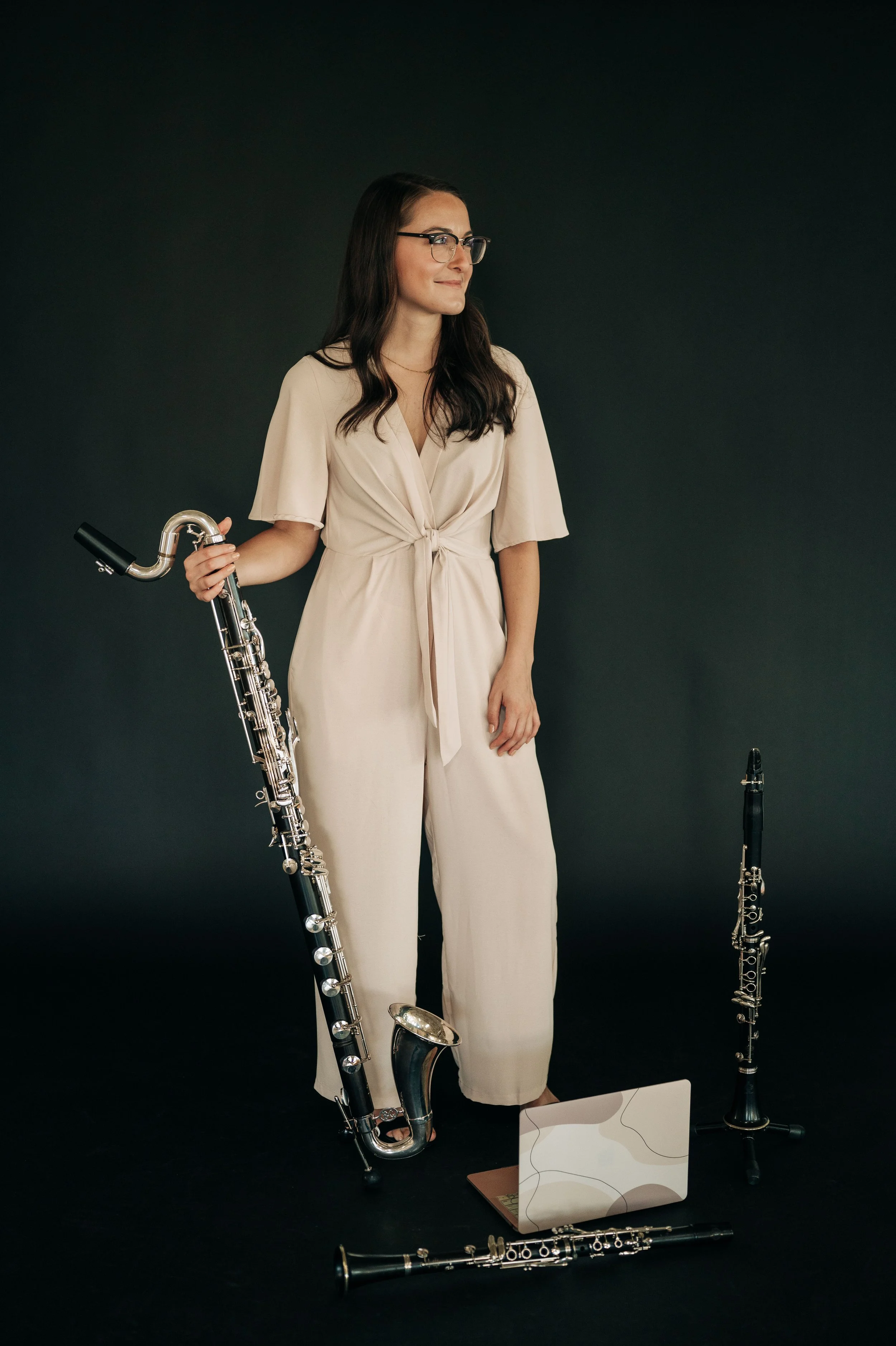 A woman in a beige jumpsuit with glasses holding a bassoon stands against a dark background, with another bassoon and a laptop on the floor.