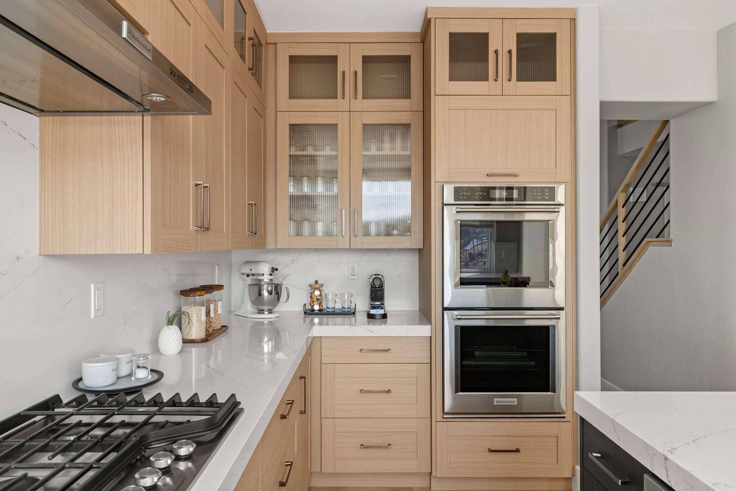 Modern kitchen with wood cabinets, white marble countertops, stainless steel appliances, and a staircase in the background.