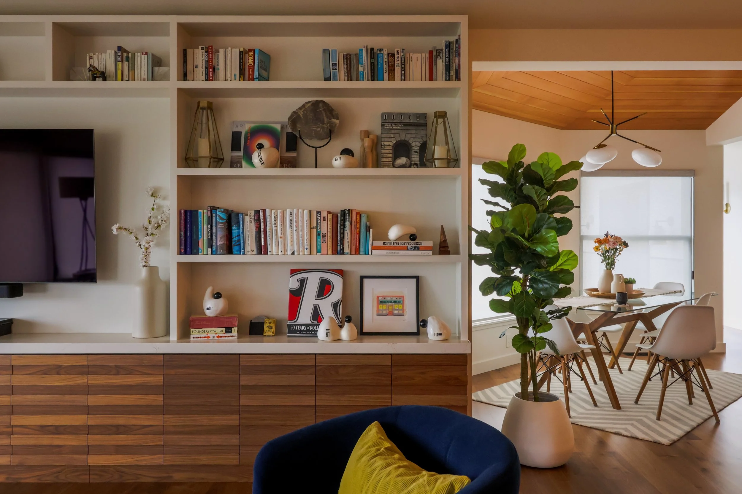 A built-in TV unit with bookshelves, blue swivel chair in the foreground, breakfast nook with a fiddle-leaf fig plant to the side.