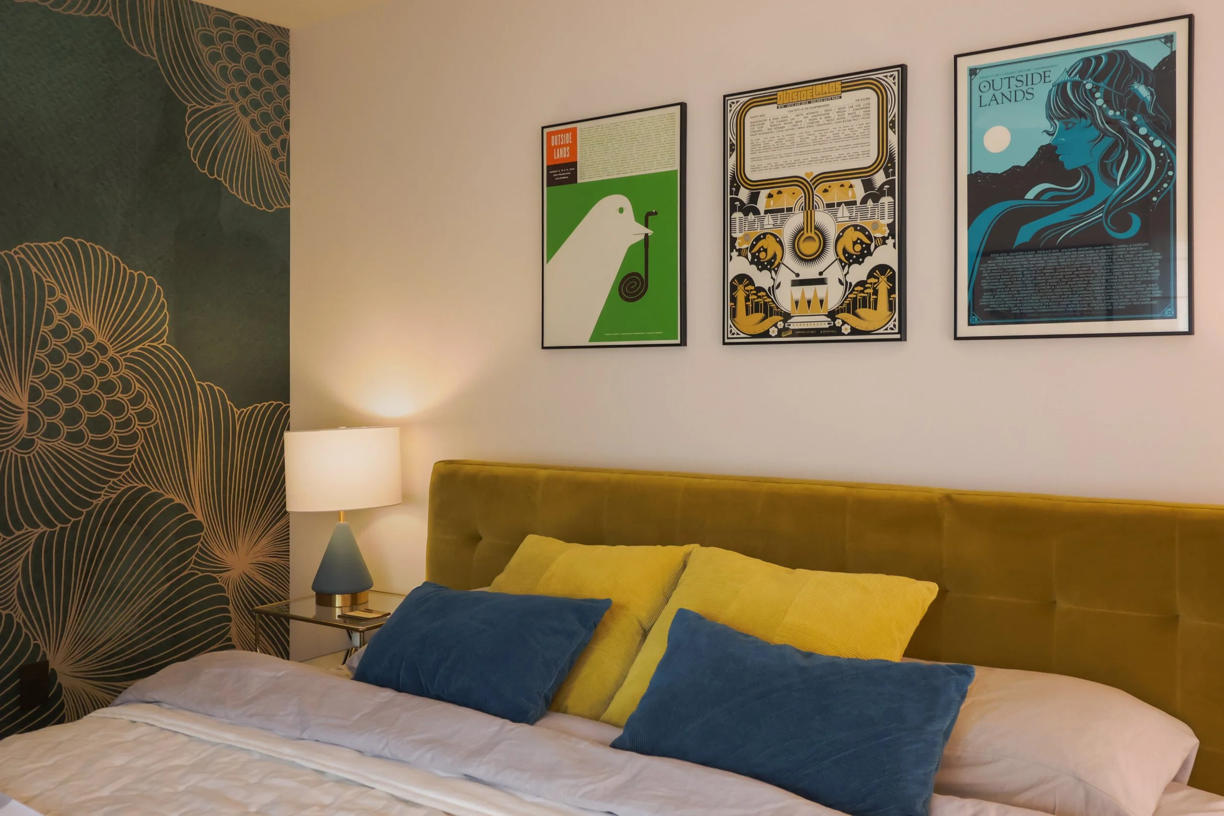 A bedroom with yellow and blue pillows on the bed, posters for outside lands are mounted above the headboard, a green and gold floral wallpaper on one of the walls.
