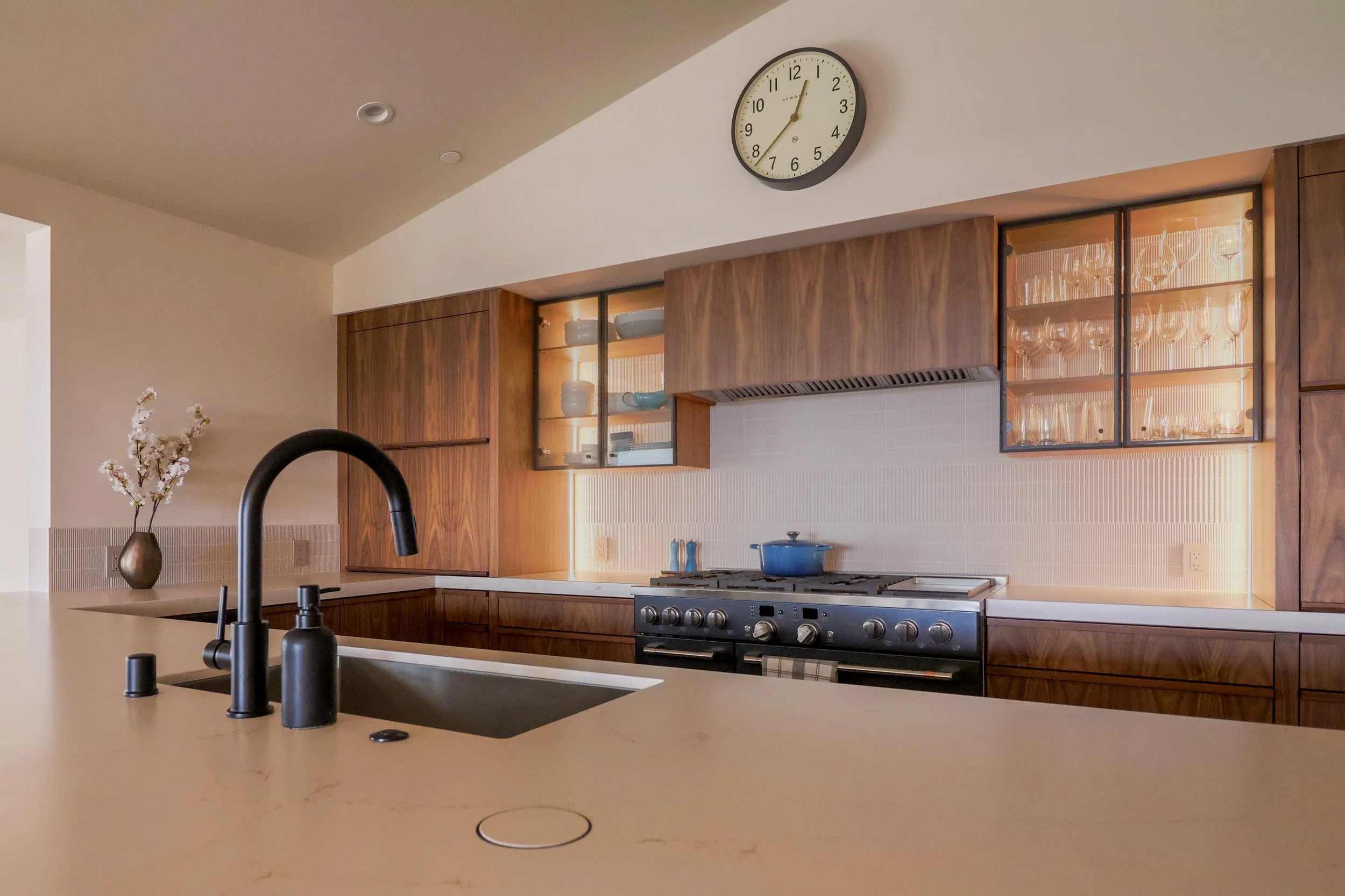 A kitchen with walnut cabinetry and warm countertops. Black faucetry and a stainless steel range are prominent, glass-fronted display cabinets flank the built-in range hood.