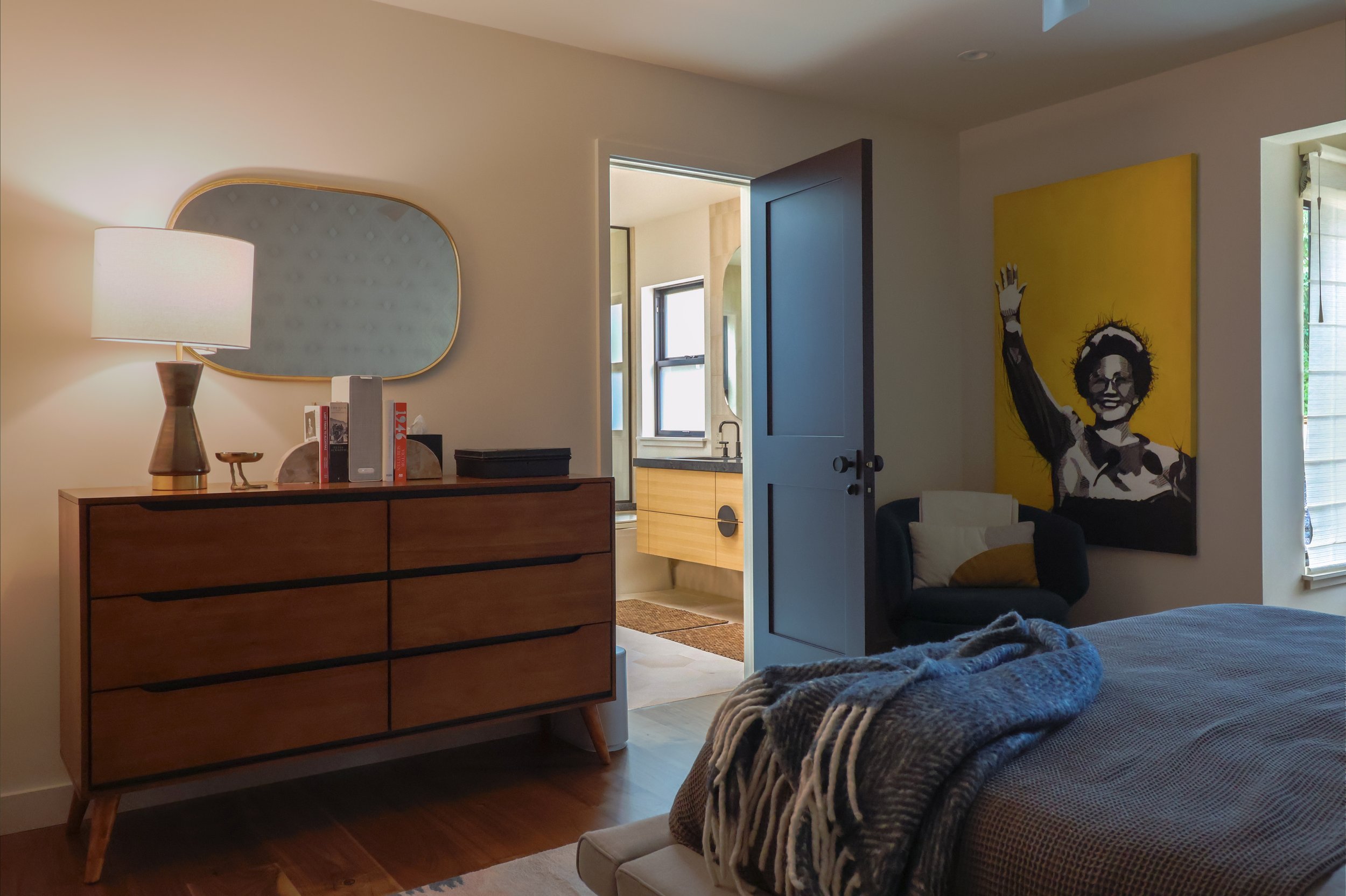 A bedroom with the foot of the bed and dresser visible. A large yellow painting fills one of the walls, a small mirror is mounted to the wall above the dresser, the door to the bathroom is open.