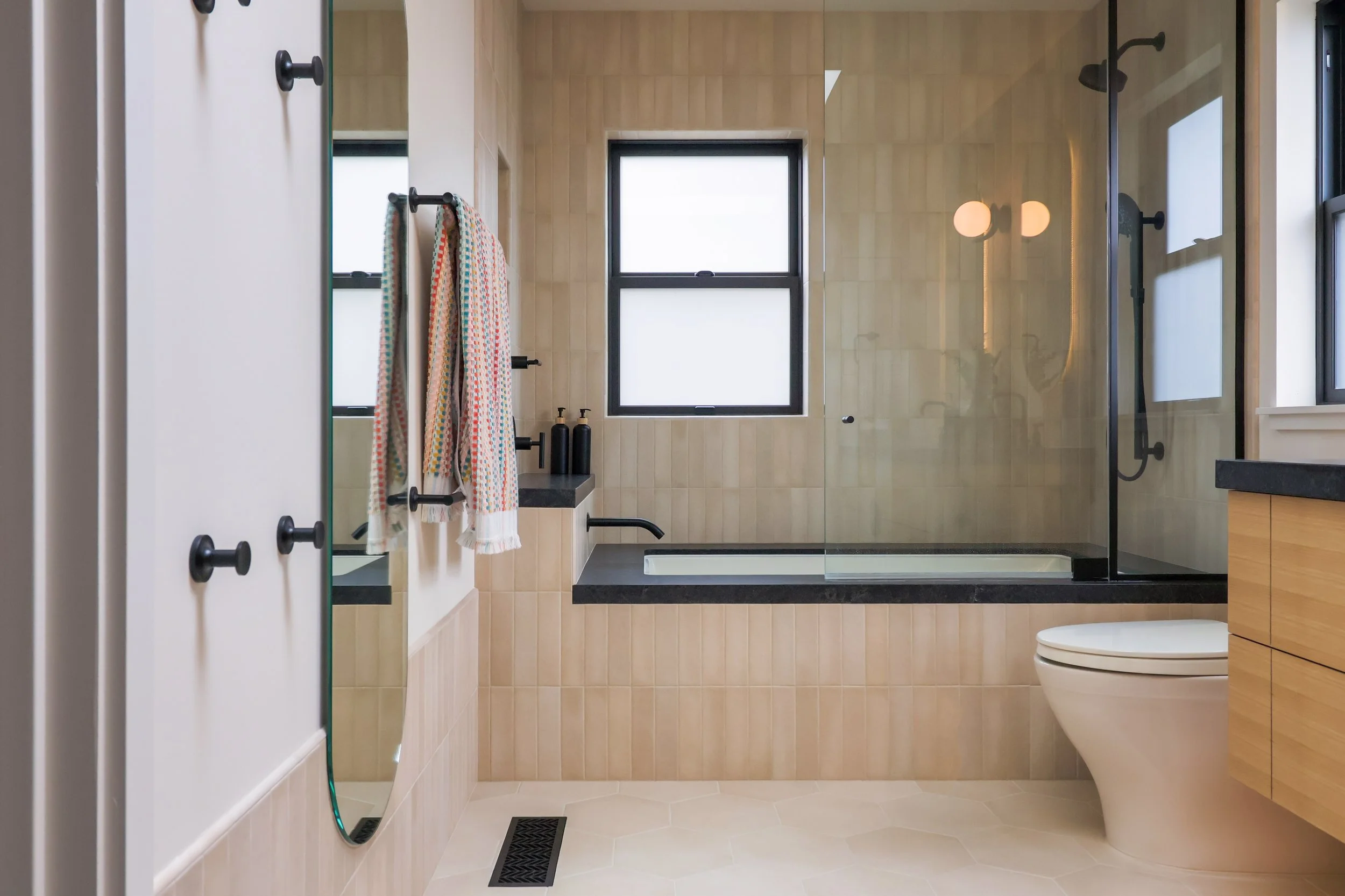 A contemporary bathroom with white oak cabinets, a dark soapstone countertop and beige tiles run all the way to the ceiling. The window over the bathtub illuminates the space, towels hang from the towel rail.