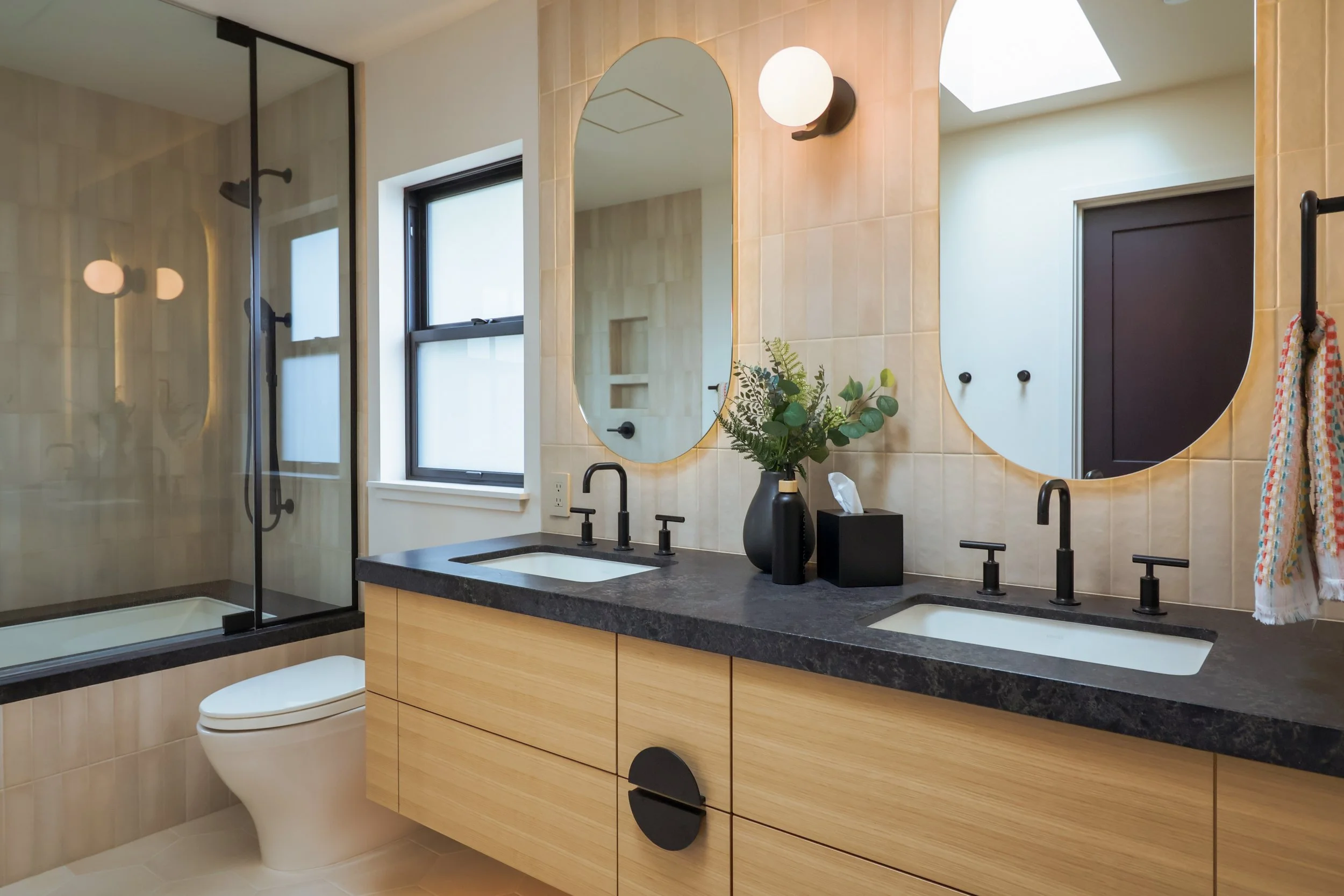 A contemporary bathroom with white oak cabinets, a dark soapstone countertop and beige tiles run all the way to the ceiling.
