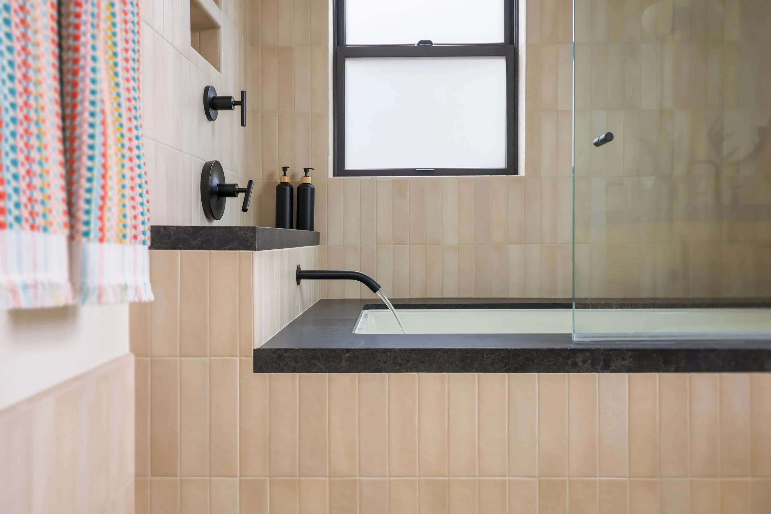 A bath tub being filled with water from a faucet, the tile is beige and contrasted by black stone and faucetry.