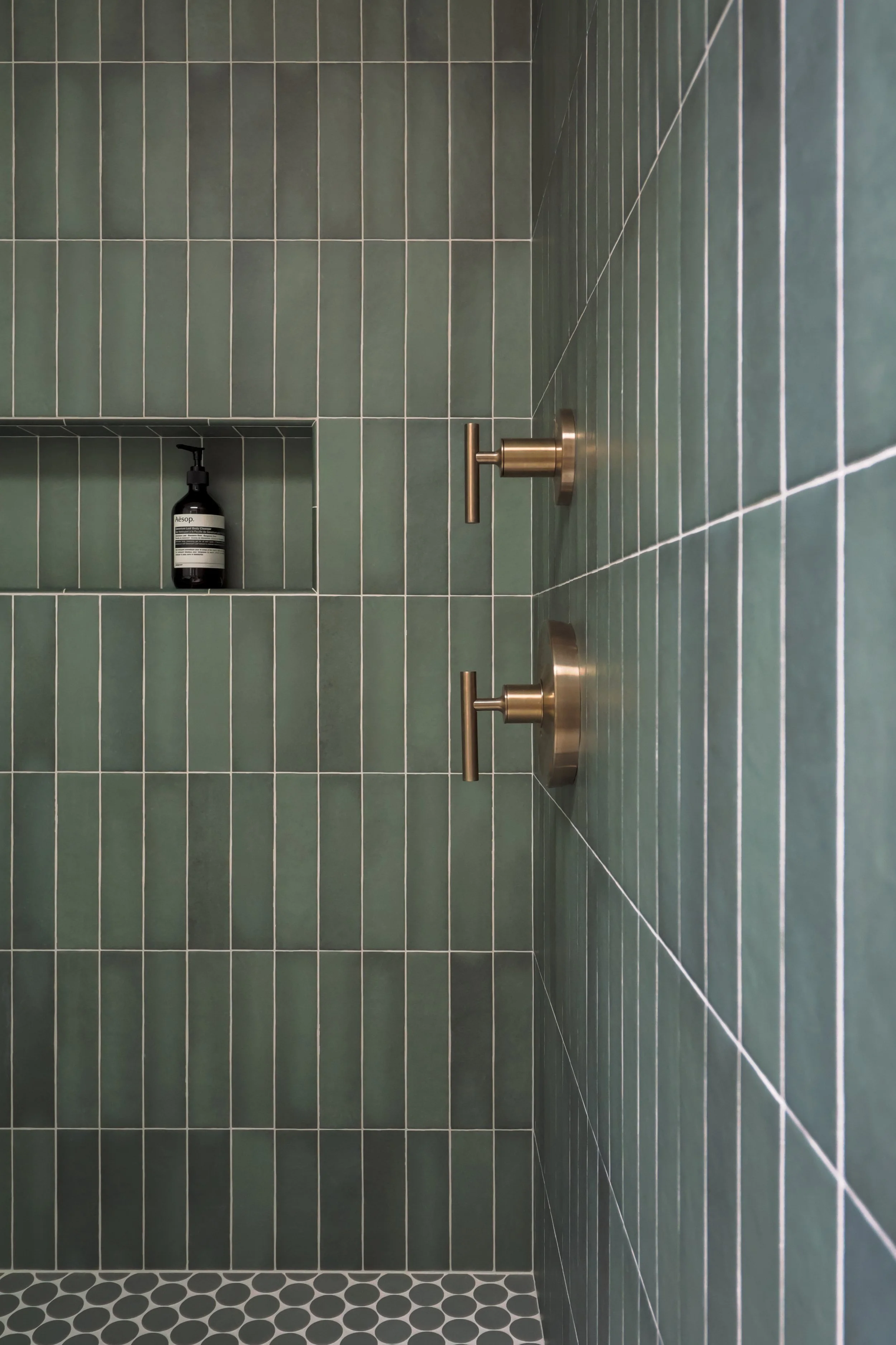 A green tiled shower stall with brushed bronze shower faucetry mounted on the wall, a shower niche holds a black shampoo bottle.