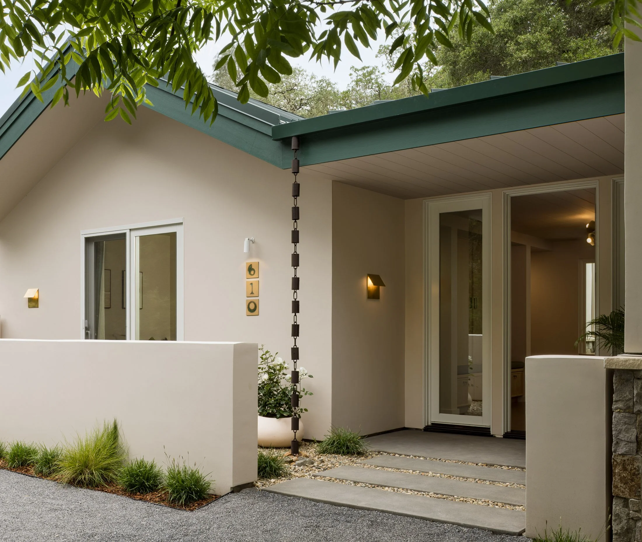 Contemporary house exterior with large windows, a green roof, and a hanging wind chime, surrounded by greenery and small plants.