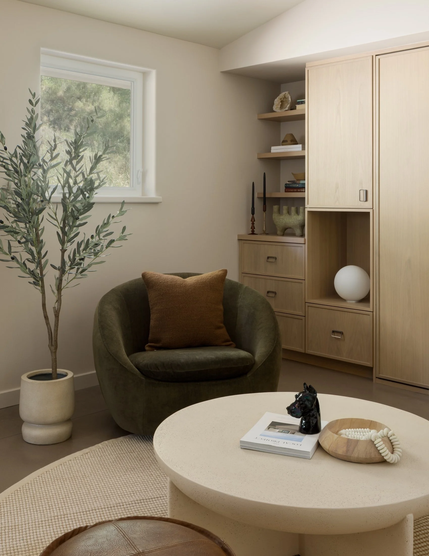 A cozy living room corner featuring a large window with a view of trees, a dark velvet armchair with a brown pillow, and a modern white coffee table with decor including a black sculpture, a book, a wooden dish with beads, and a white sphere. A potte
