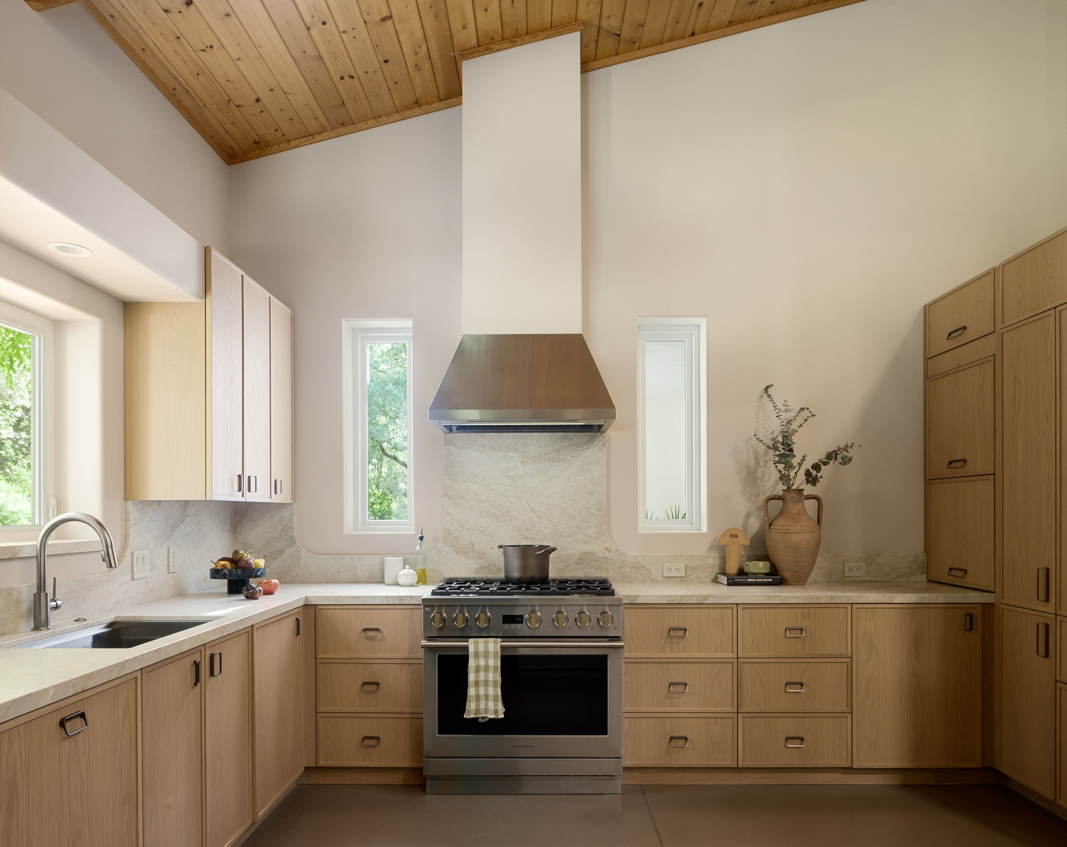 Contemporary kitchen with beige cabinets, stainless steel stove, wooden vaulted ceiling, and two small windows with greenery outside.