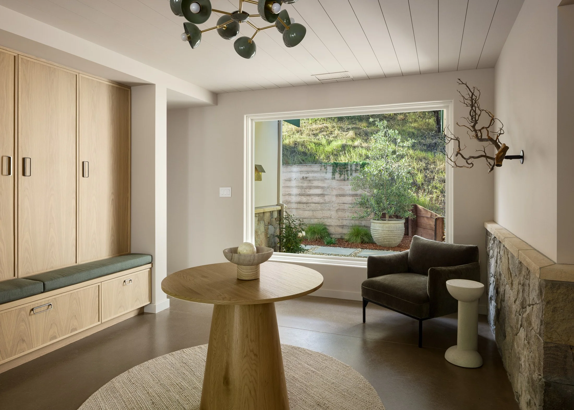 Contemporary foyer with large window, armchair, wooden dining table with a bowl, built-in wooden storage with cushion seating, decorative branch on wall, stone fireplace, plants outside visible through the window, modern lighting fixture on ceiling.