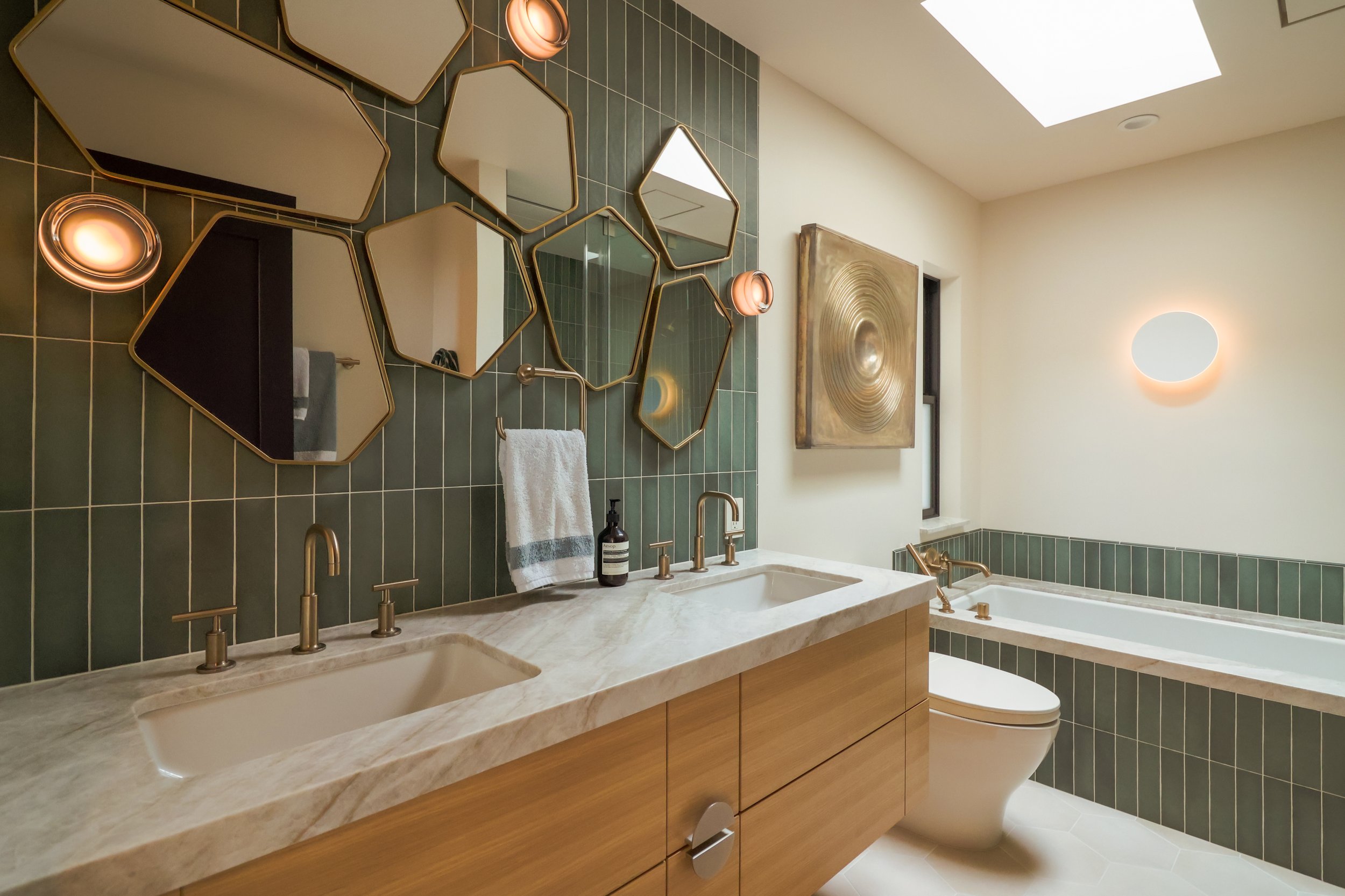 A green tiled bathroom with white oak cabinetry and taj mahal quartzite countertops. The faucetry is a brushed bronze finish which matched a 3D piece of artwork mounted above the toilet. Abstract shaped mirrors are mounted above the vanity.