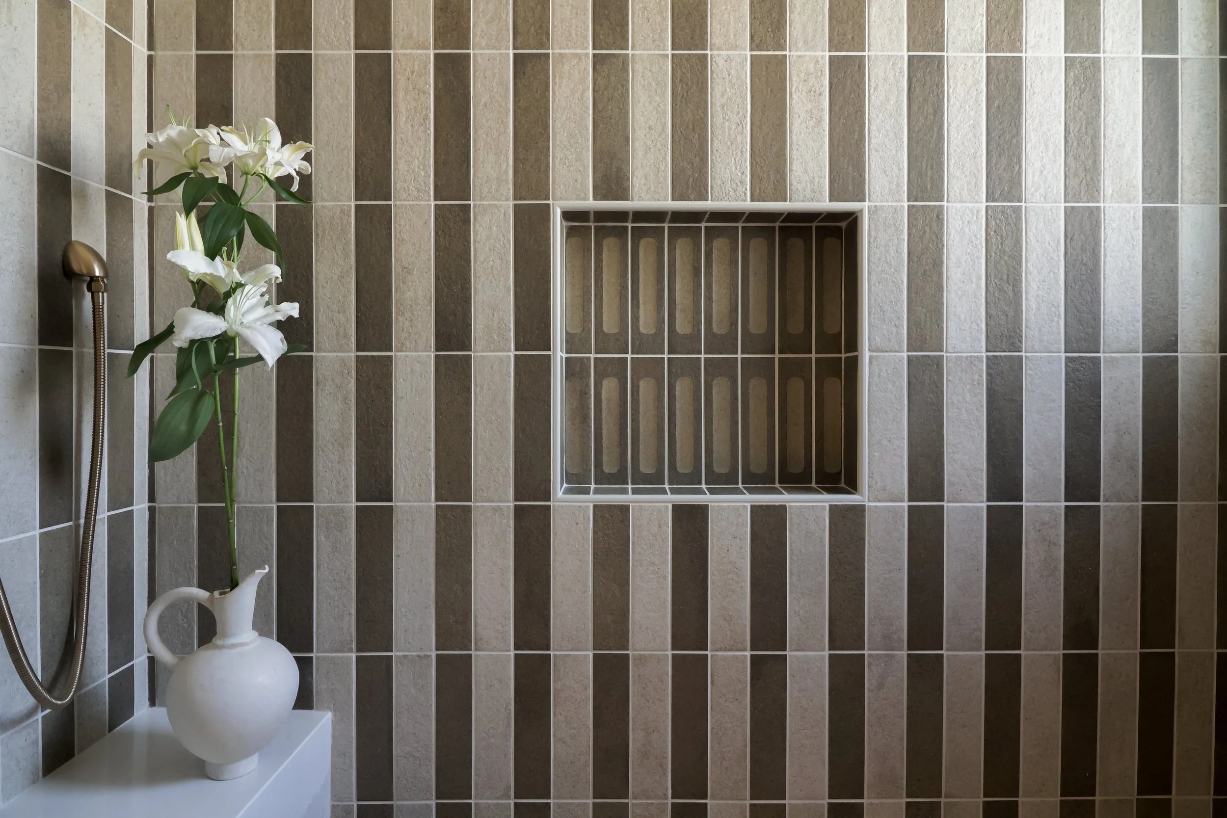 A tiled shower with alternating vertical stripes of tan and green tiles. A pitcher of white lilies sits on a shower bench on the left.