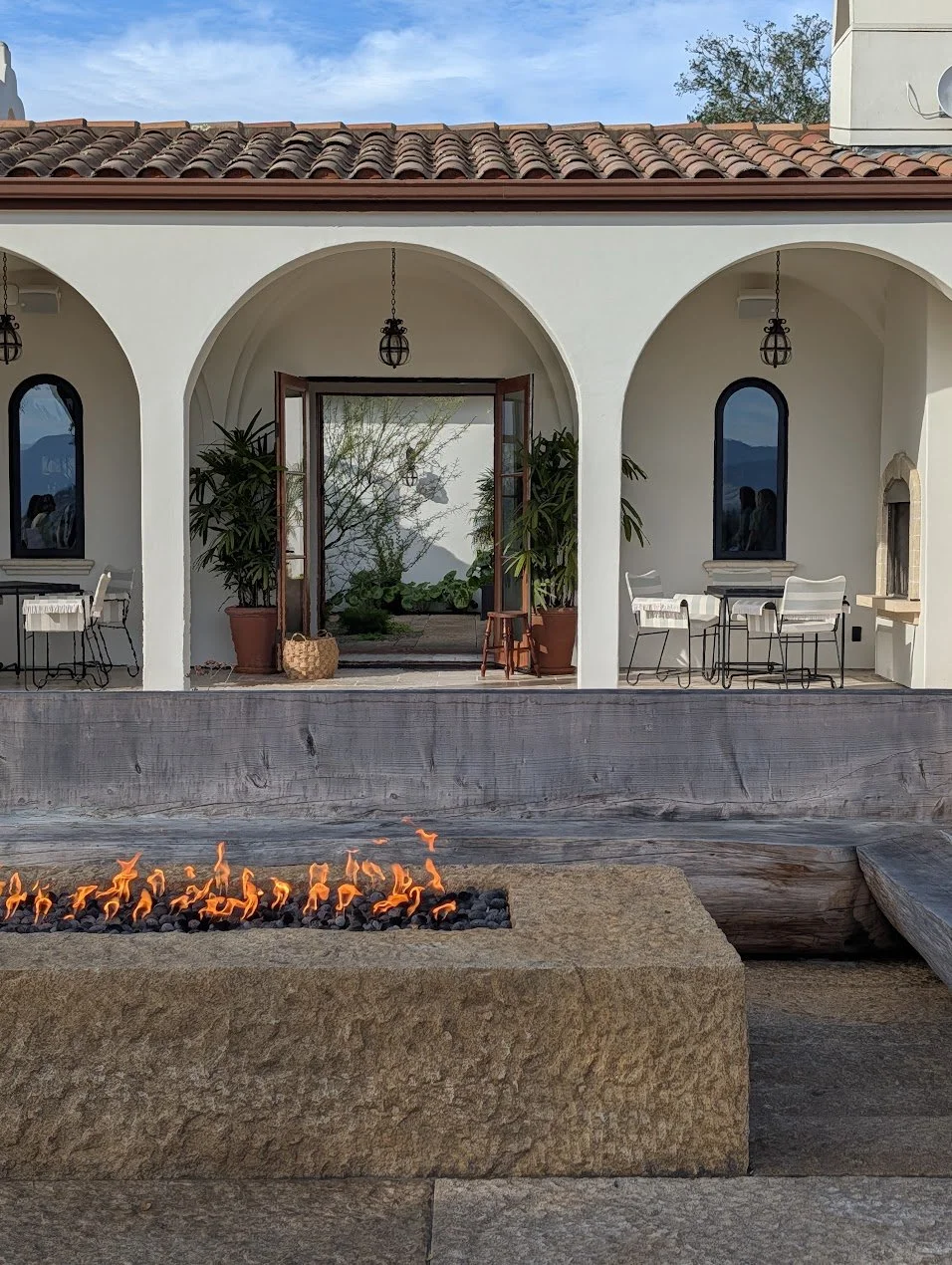 The exterior of a mission-style home with terracotta roof tiles and stucco walls with arches openings. A firepit in the foreground.