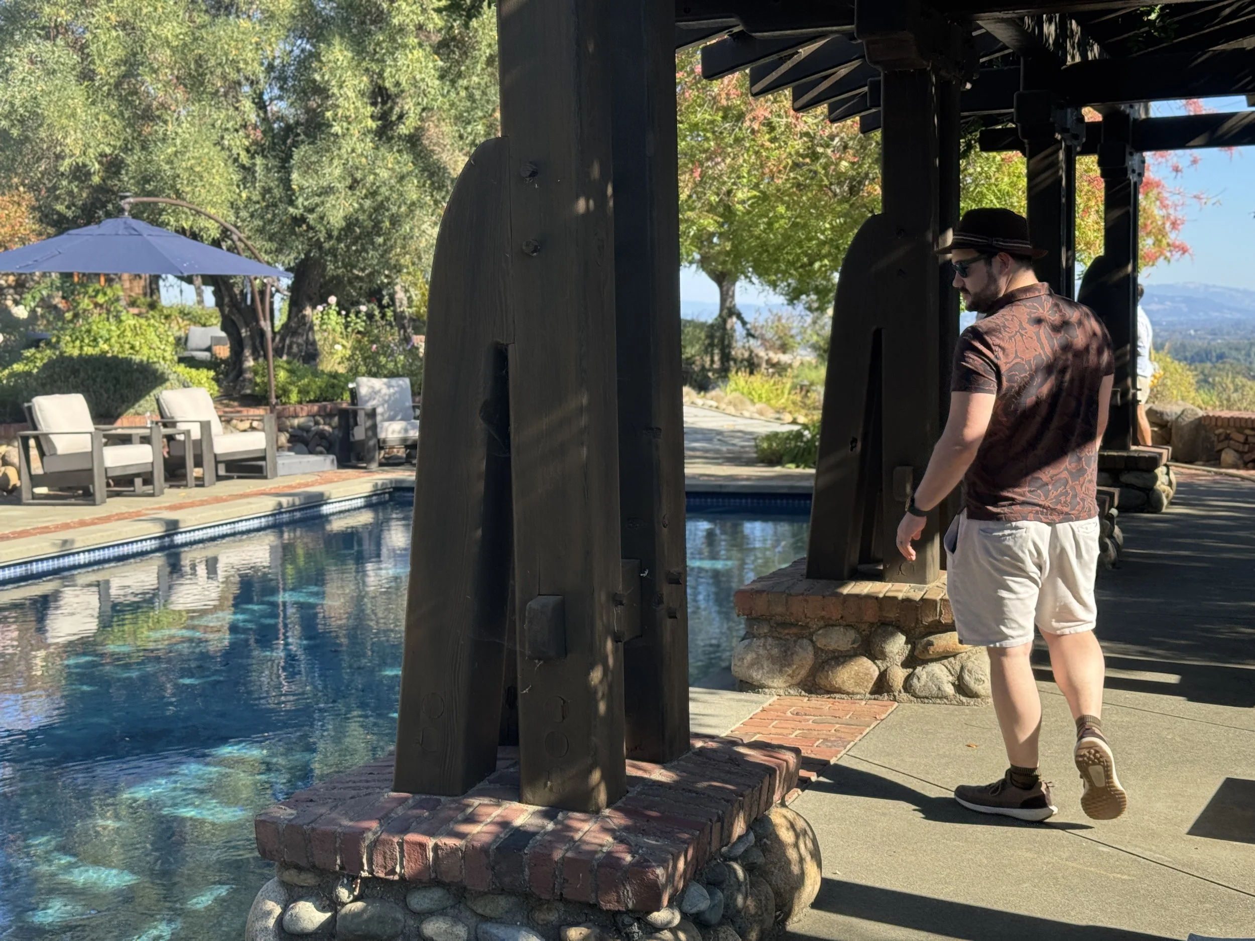 Large wooden posts support a large wooden pergola adjacent to a swimming pool.