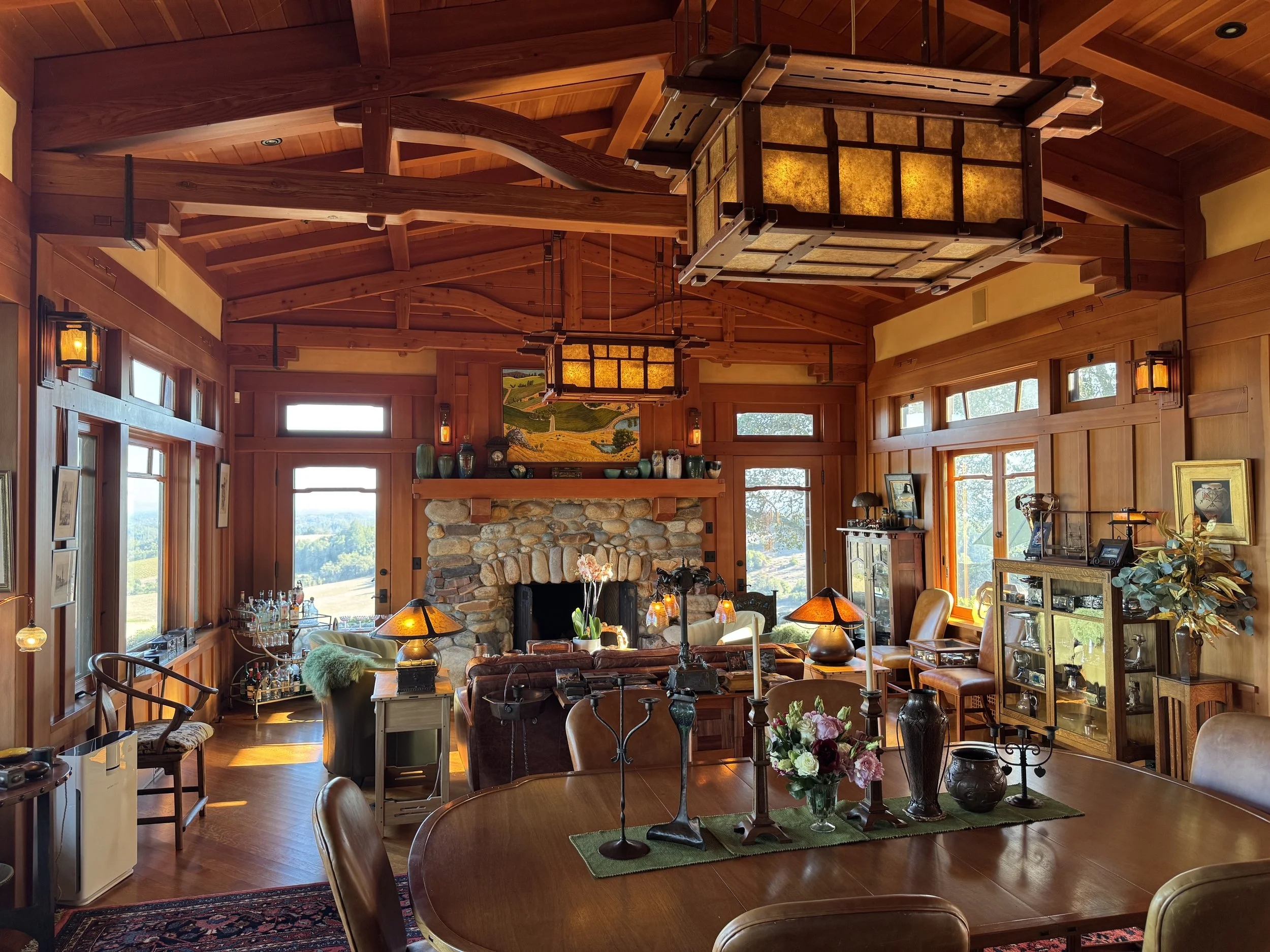 An open plan living and dining room with wood panelling and ceiling, large decorative wood beams support the roof overhead, bespoke wooden chandeliers hang over the dining and living room. Countless Arts & Crafts movement objects adorn the space.