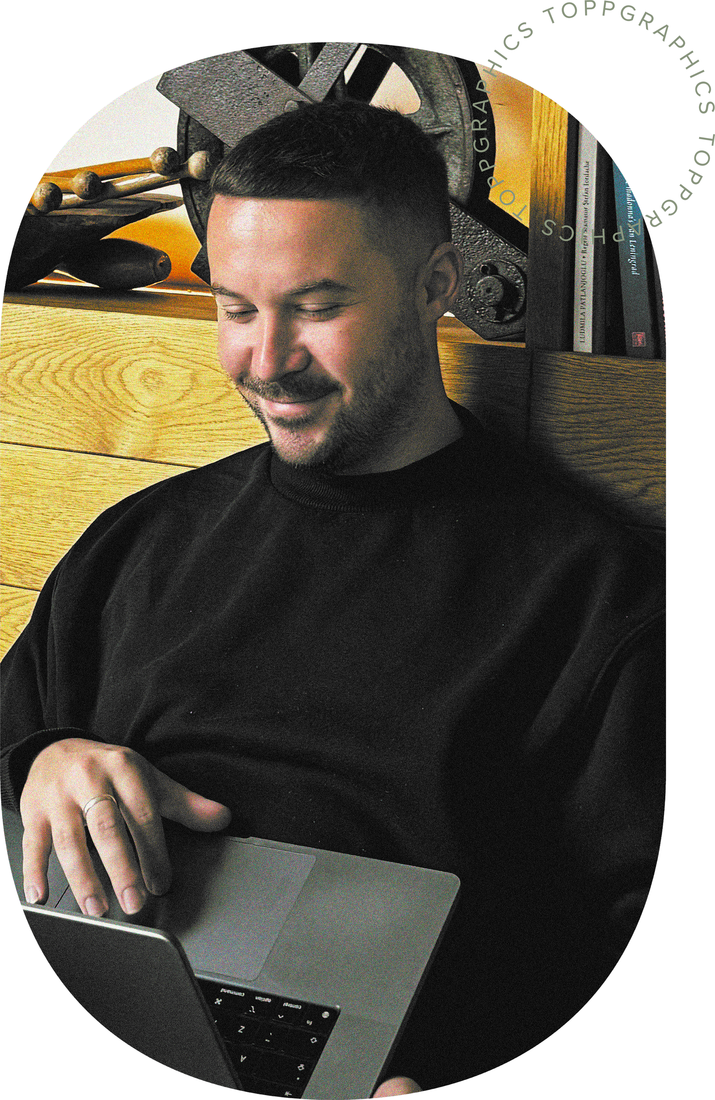 A man with short dark hair and a beard is sitting at a desk, smiling while using a laptop. He is wearing a black sweatshirt. Behind him, there is a wooden wall with a shelf holding books and a large piece of machinery or equipment.
