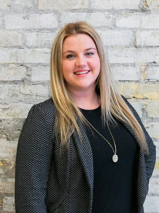 A smiling woman with long blonde hair wearing a black top and a black patterned blazer, standing in front of a white brick wall.
