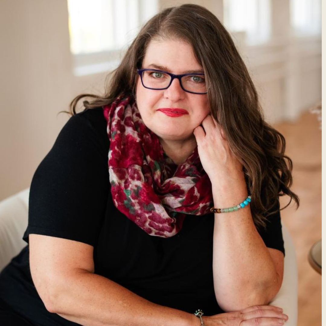 A woman with brown hair, glasses, and red lipstick is sitting indoors, resting her face on her hand, wearing a black top, a colorful scarf, and beaded bracelets.
