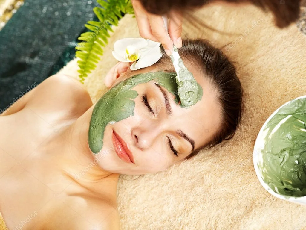 A woman is receiving a facial treatment with green clay mask applied on her face, lying on a massage table with her eyes closed, surrounded by spa elements like a white orchid flower and green ferns.