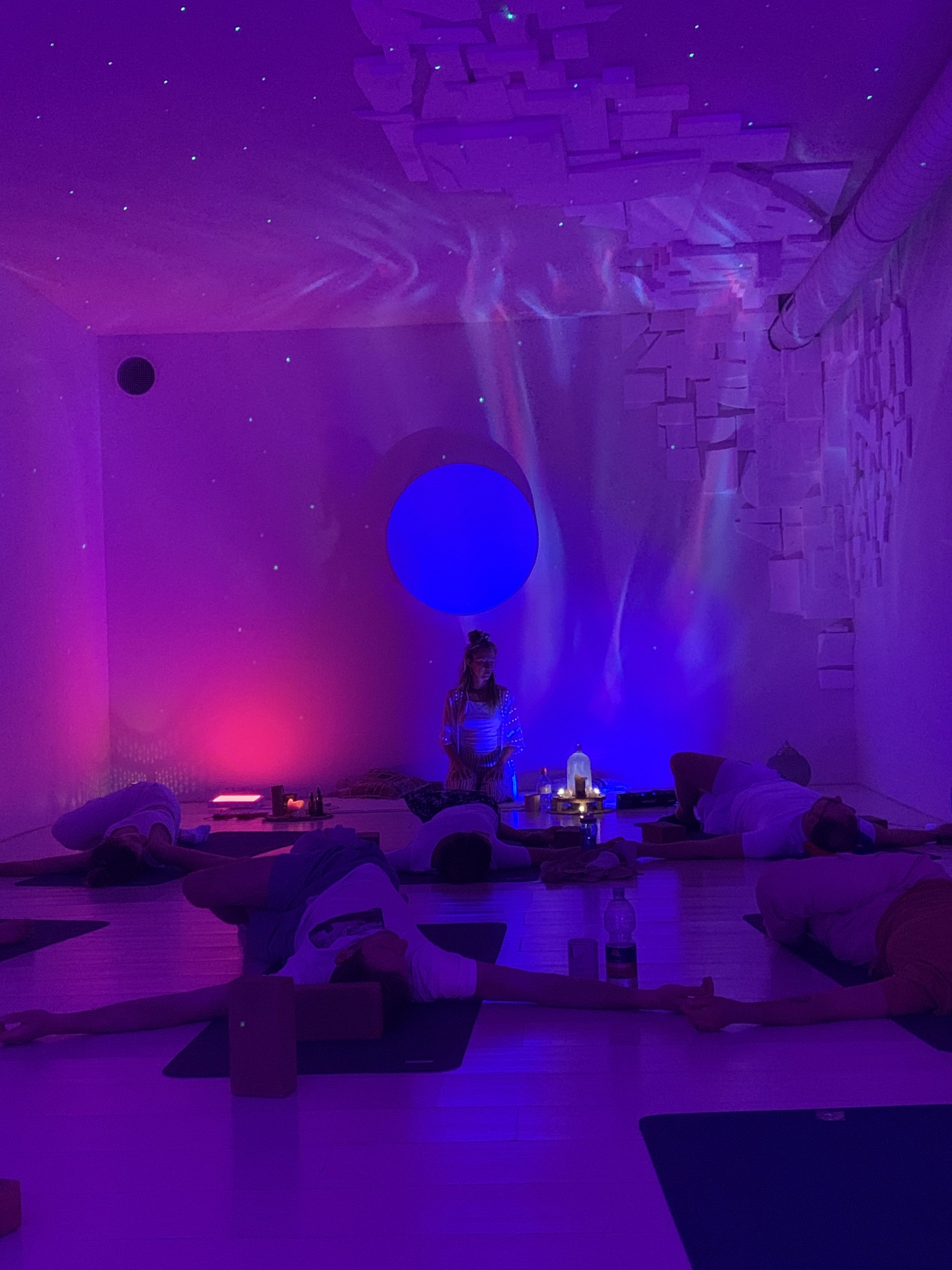 Personen praktizieren Yoga in einem Raum mit bunter Beleuchtung und entspannender Atmosphäre.