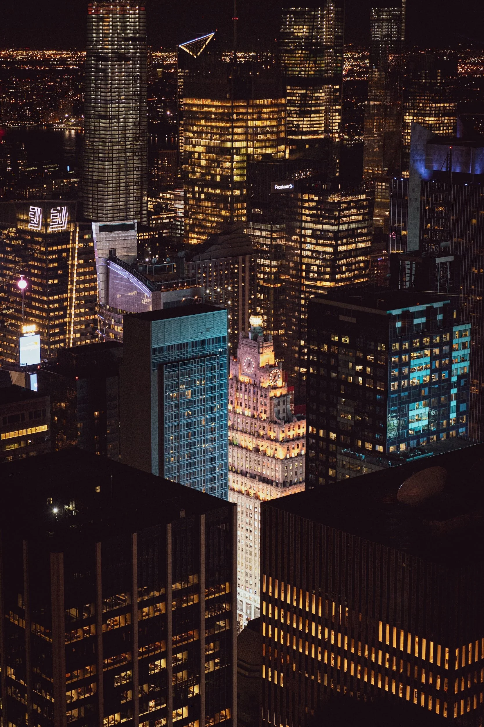 Nighttime view of a city skyline with illuminated skyscrapers, including the clock tower of the New York Hilton Hotel, surrounded by other tall office buildings with lights on.