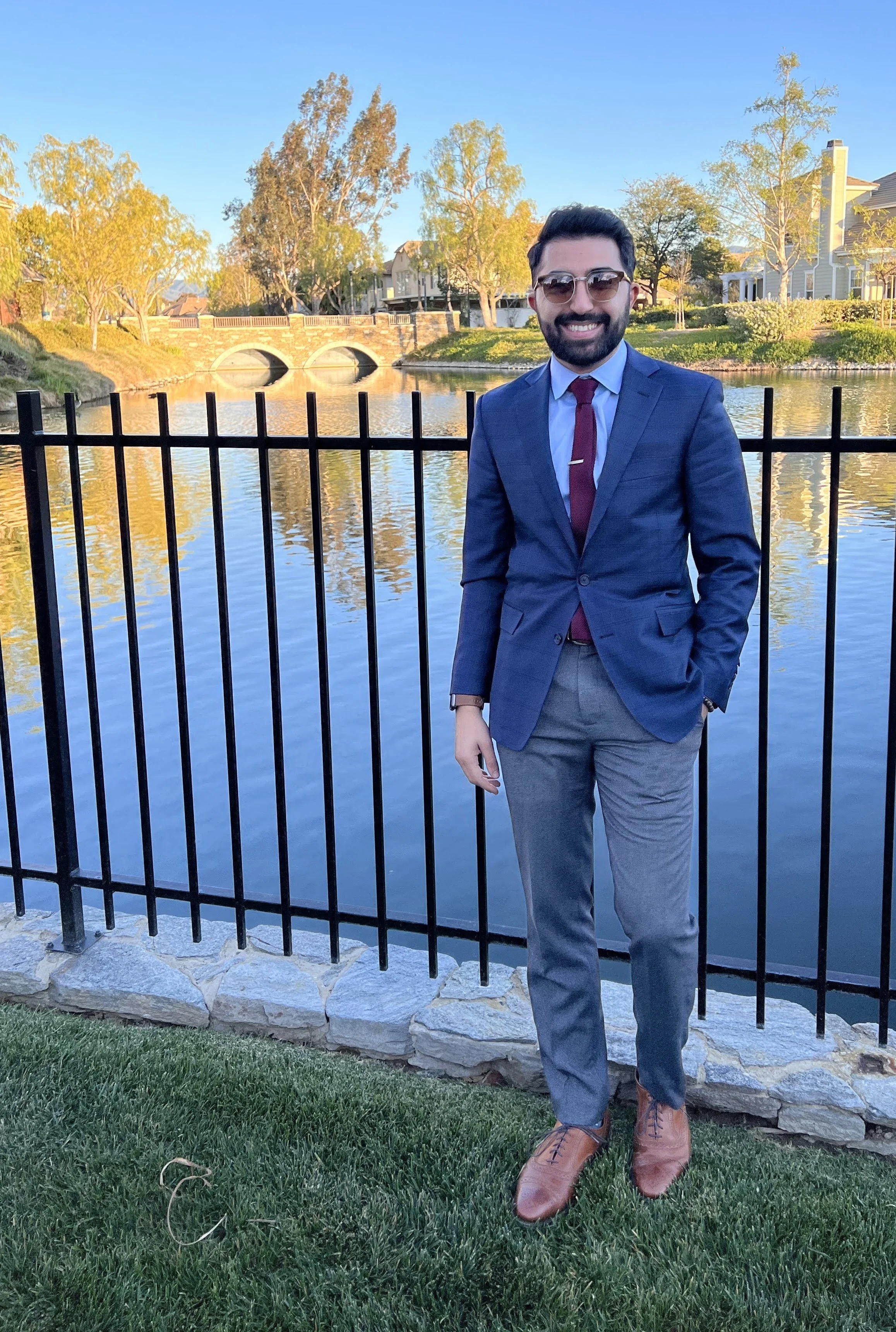A man in a blue suit and tie, wearing glasses, standing outdoors by a black metal fence near a lake with trees and houses in the background during golden hour.
