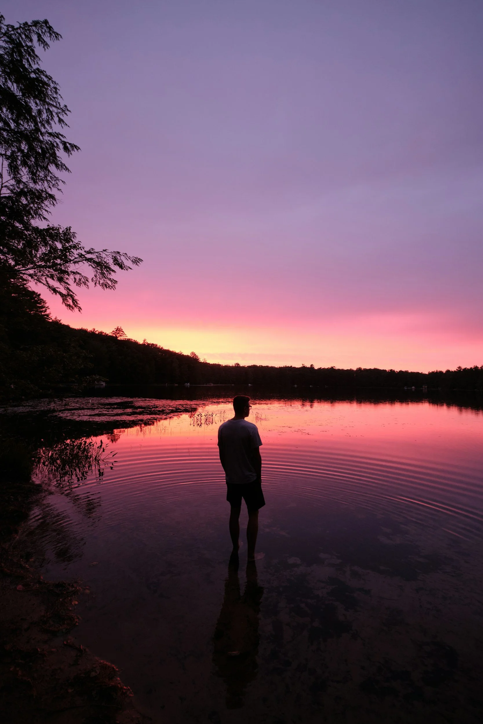 Silhouette of a person with long hair standing near water during a colorful sunset with pink, orange, and purple clouds.