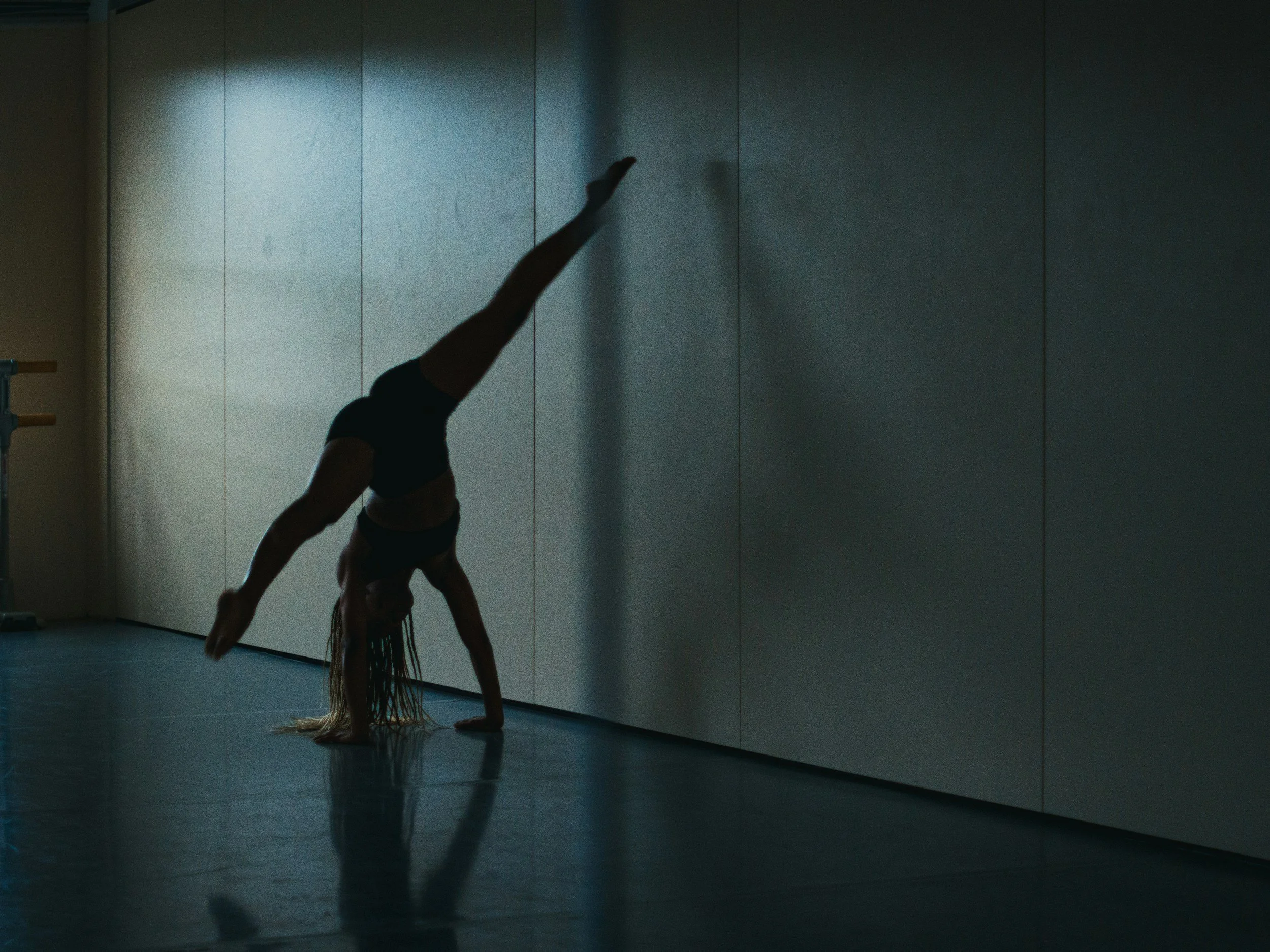 A woman performing a handstand in a gym or dance studio, with one leg extended upwards and the other bent, against a plain wall with minimal lighting.