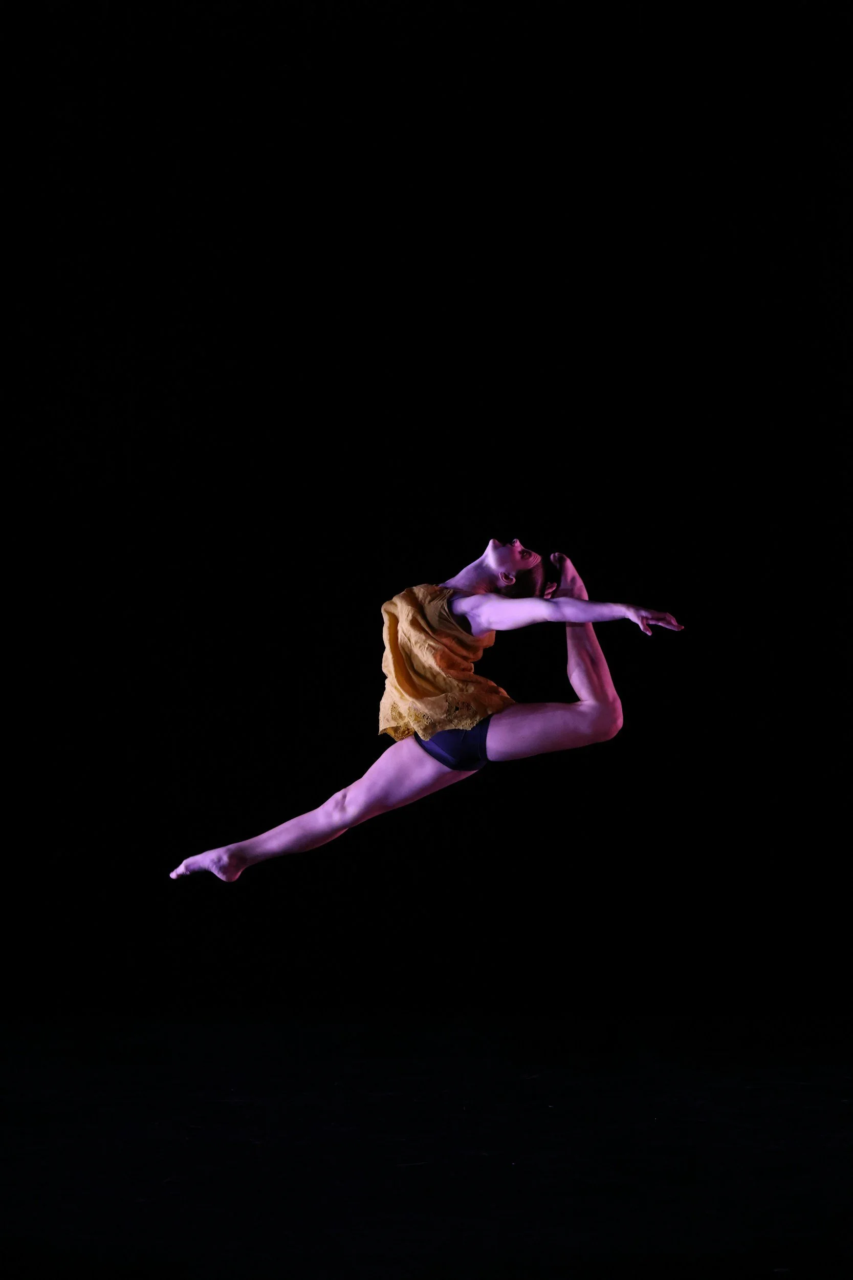 A female dancer captured in mid-air leap against a black background, wearing a yellow top and black shorts.