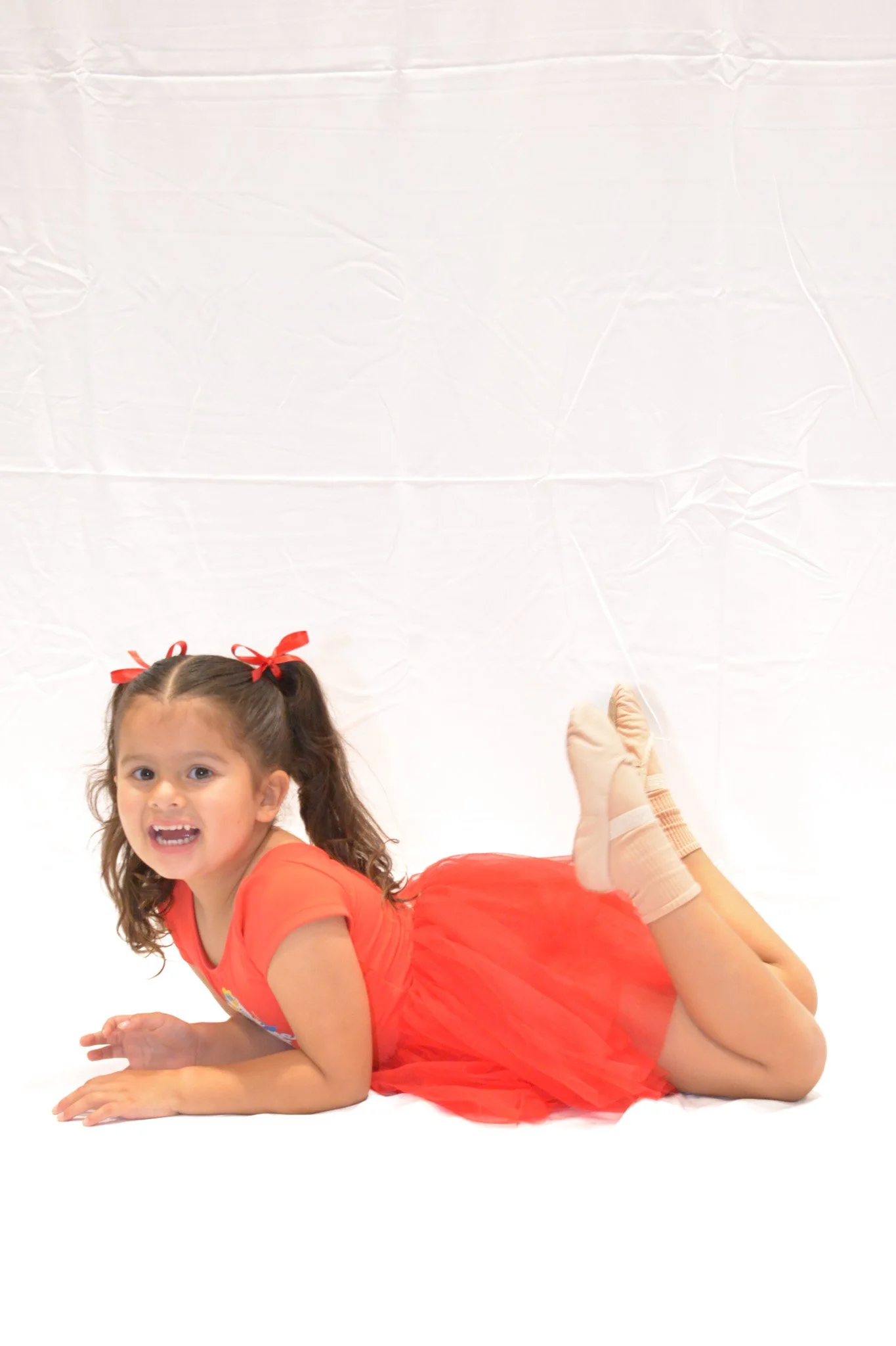 A young girl lying on her stomach in a red dress and beige socks, smiling at the camera, with her legs bent at the knees and feet crossed.