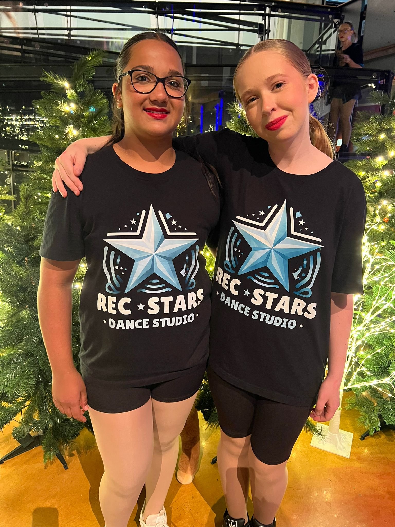 Two young women in black T-shirts with 'REC STARS DANCE STUDIO' logo standing in front of Christmas trees, smiling and posing for the camera.