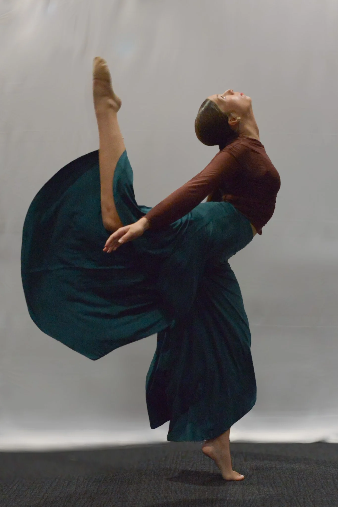 A woman performing a dance pose, balancing on one foot with the other leg raised high behind her, and her back arched with her head tilted back.