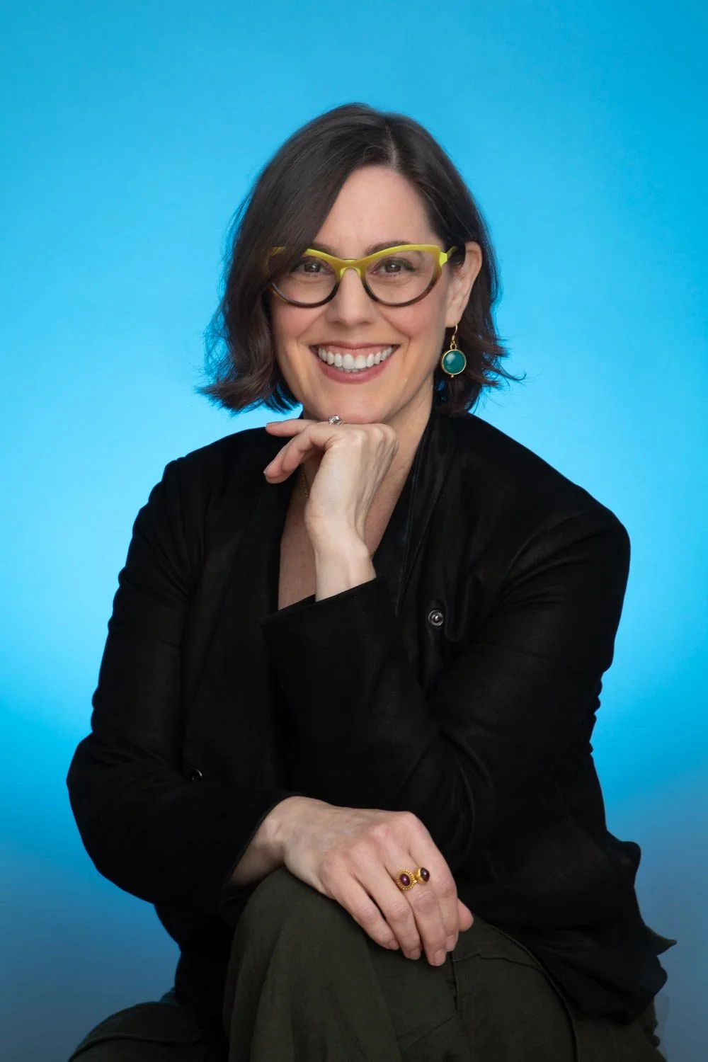Smiling, dark haired woman wearing green glasses and large dangling earrings in front of blue background