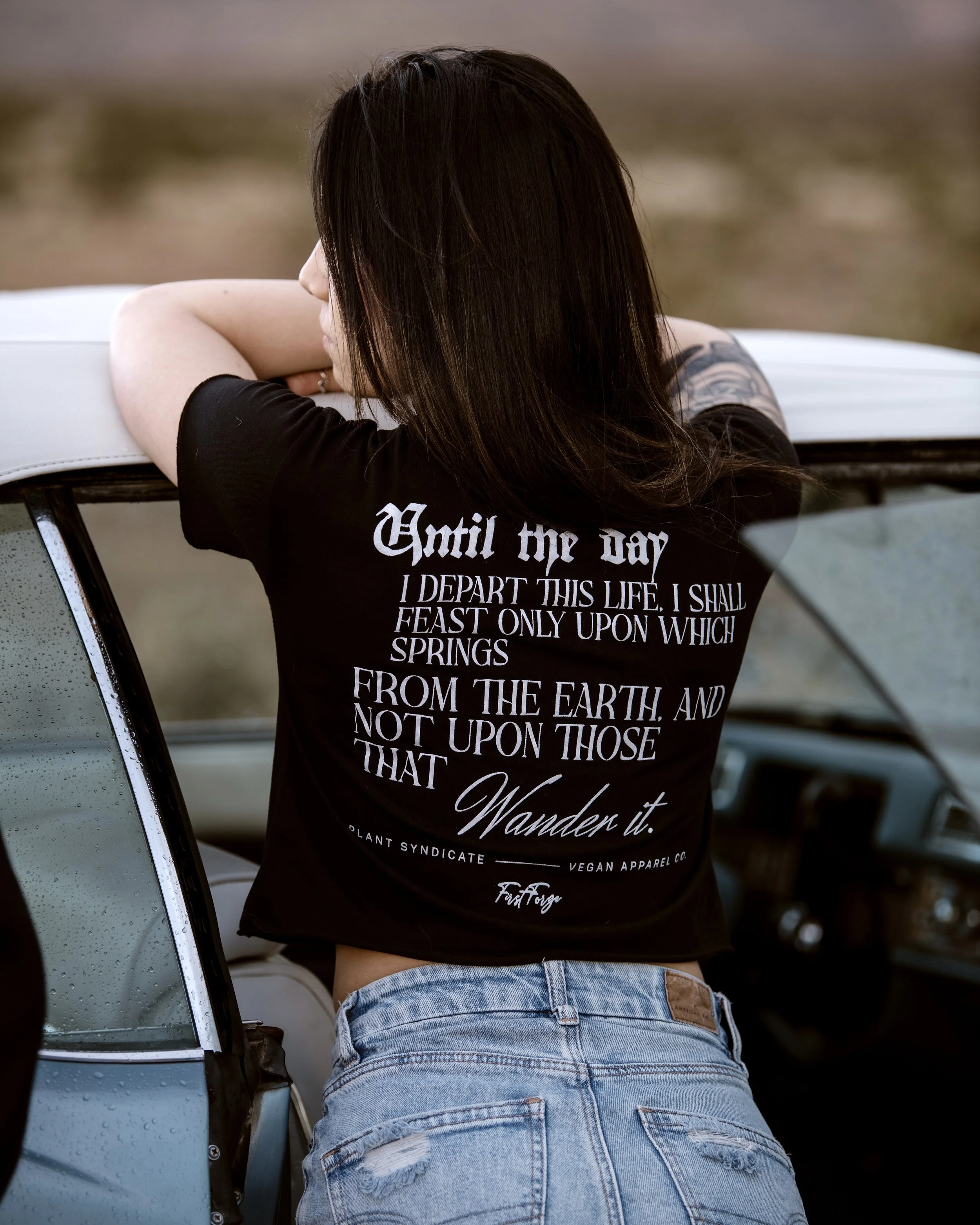 Model wearing Plant Syndicate "Until the Day" vegan apparel tee leaning on classic car, showcasing poetic back print — ethical streetwear photography.