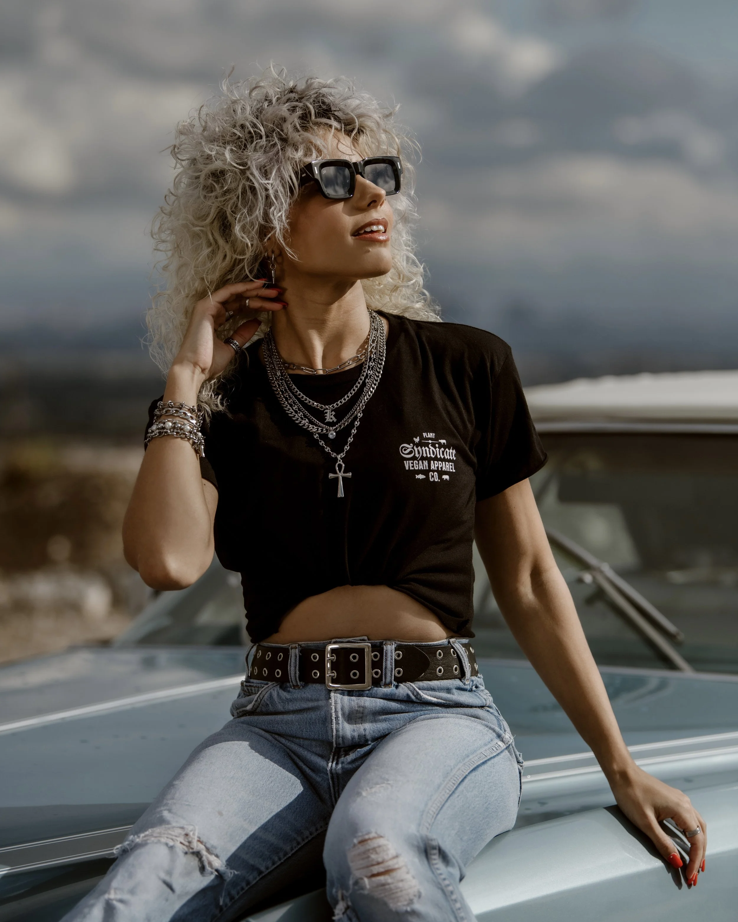 Model wearing Plant Syndicate vegan apparel black tee and layered chains, sitting on classic car under dramatic sky — ethical streetwear fashion.