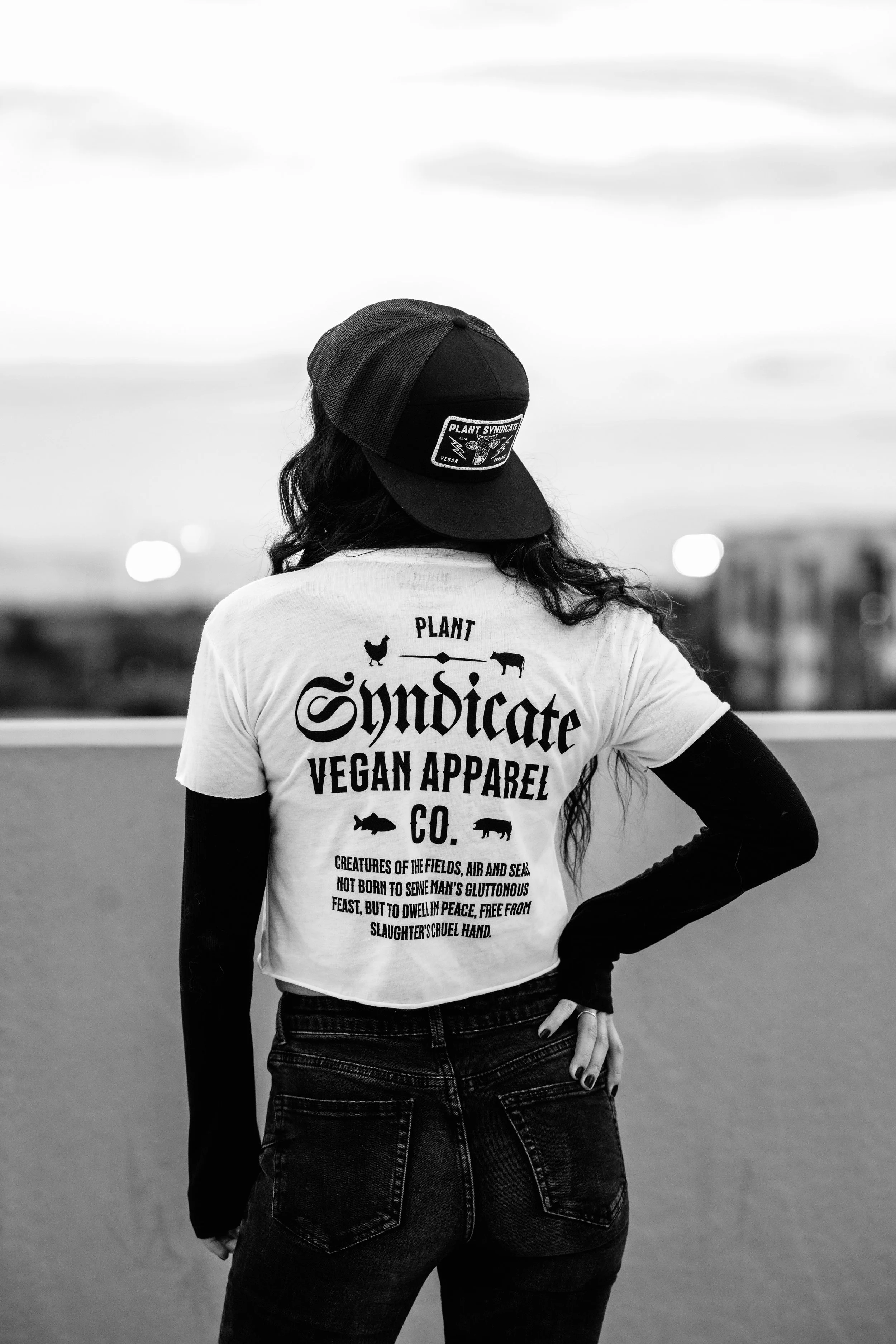 Black-and-white photo of model wearing Plant Syndicate vegan apparel and trucker hat, standing on rooftop at sunset — ethical streetwear brand.