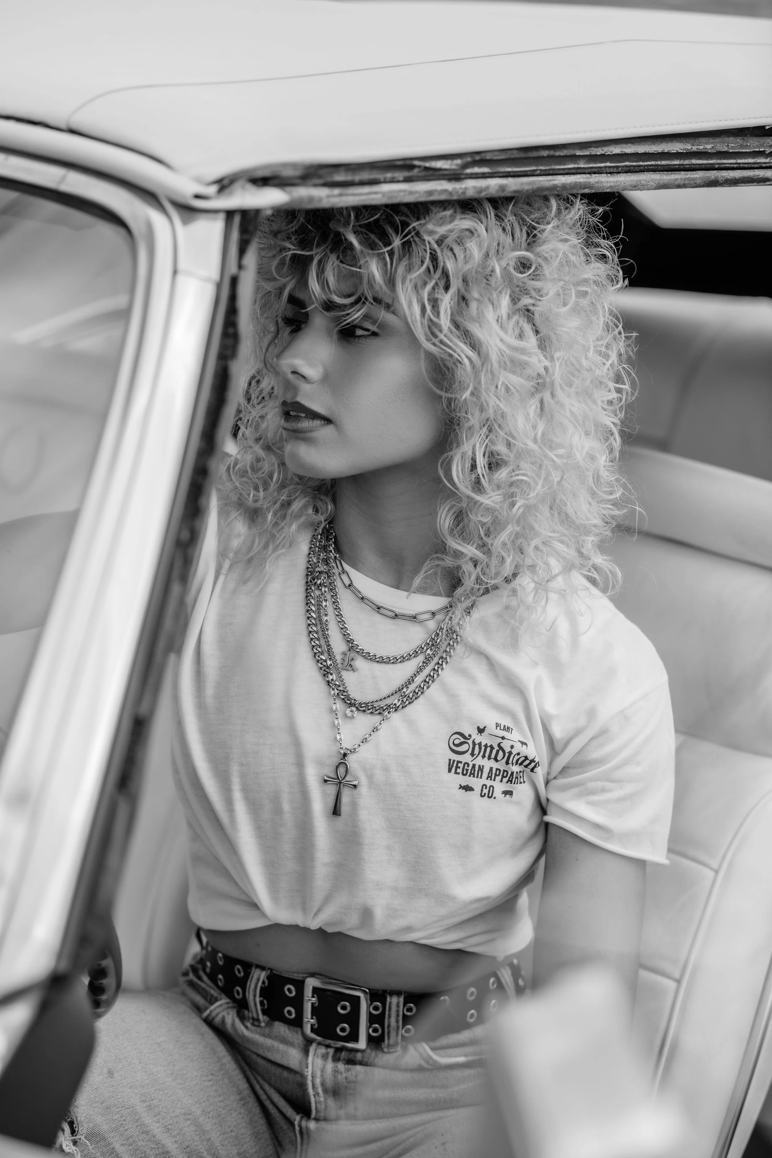 Black-and-white portrait of a curly-haired model wearing Plant Syndicate vegan apparel inside a vintage car — ethical streetwear style.
