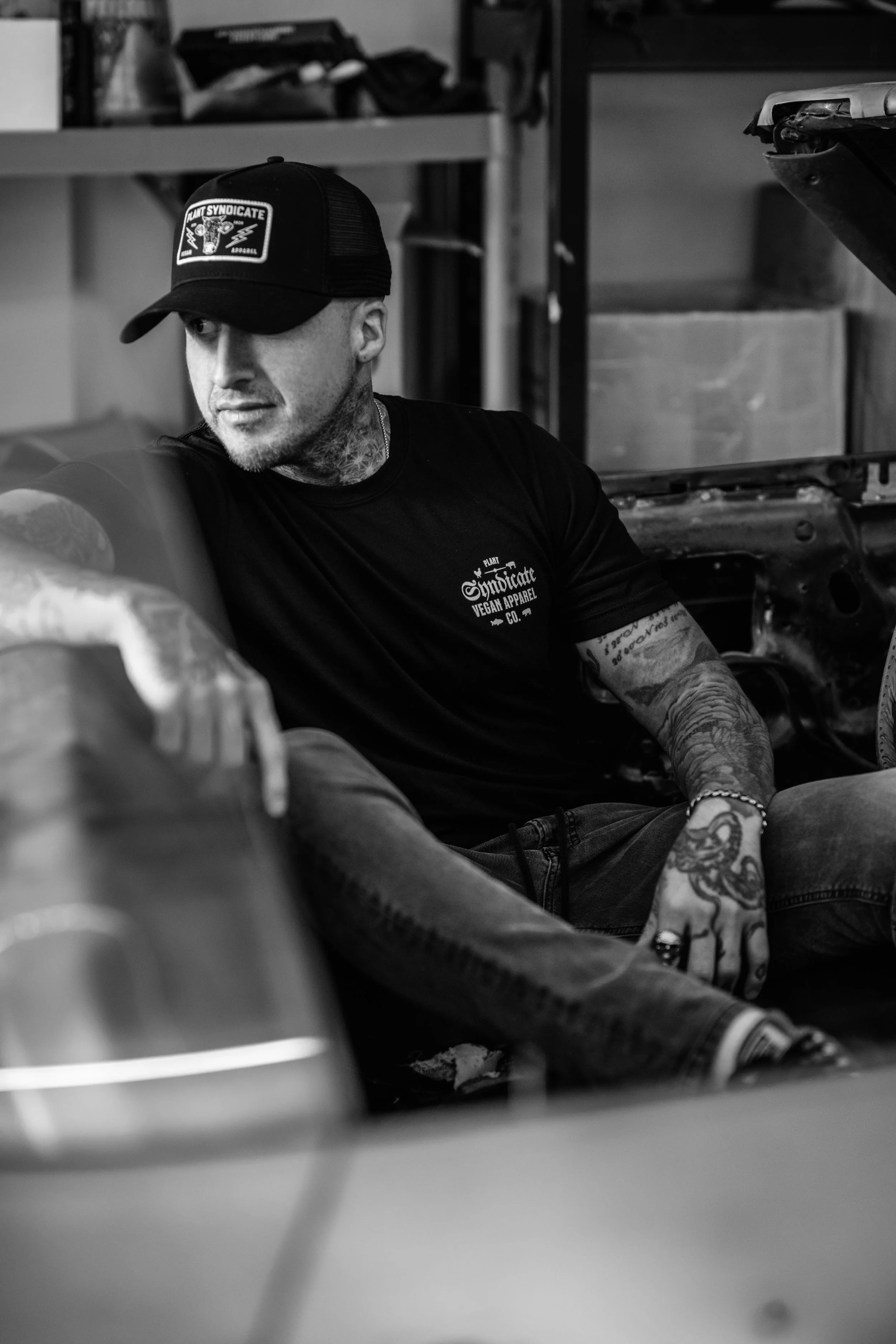 Tattooed model wearing a Plant Syndicate vegan apparel and trucker hat, sitting in a vintage car during a black-and-white industrial photoshoot — ethical vegan streetwear aesthetic.