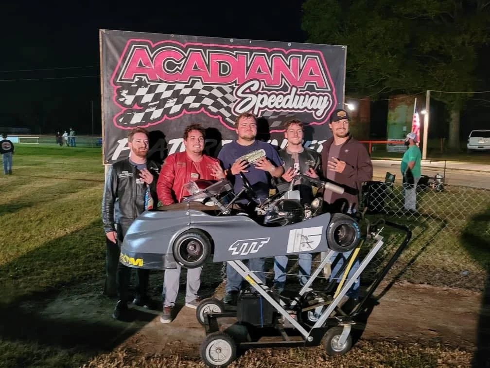 Five men standing in front of a large ACADANA Speedway sign, with a small race car and a racing simulator in front of them, at night.