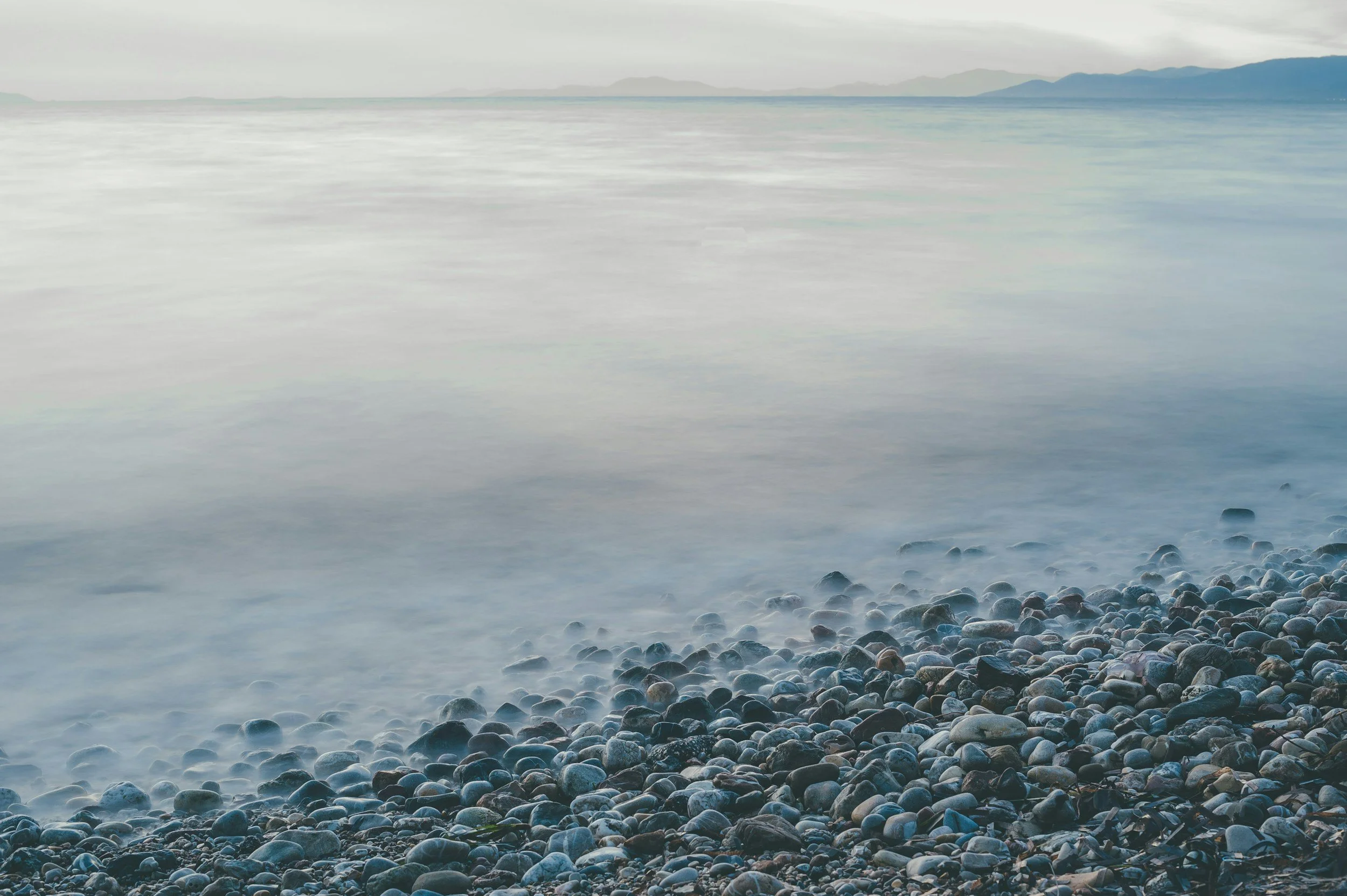 Calm sea with a rocky shore in the foreground, overcast sky, distant mountains on the horizon.