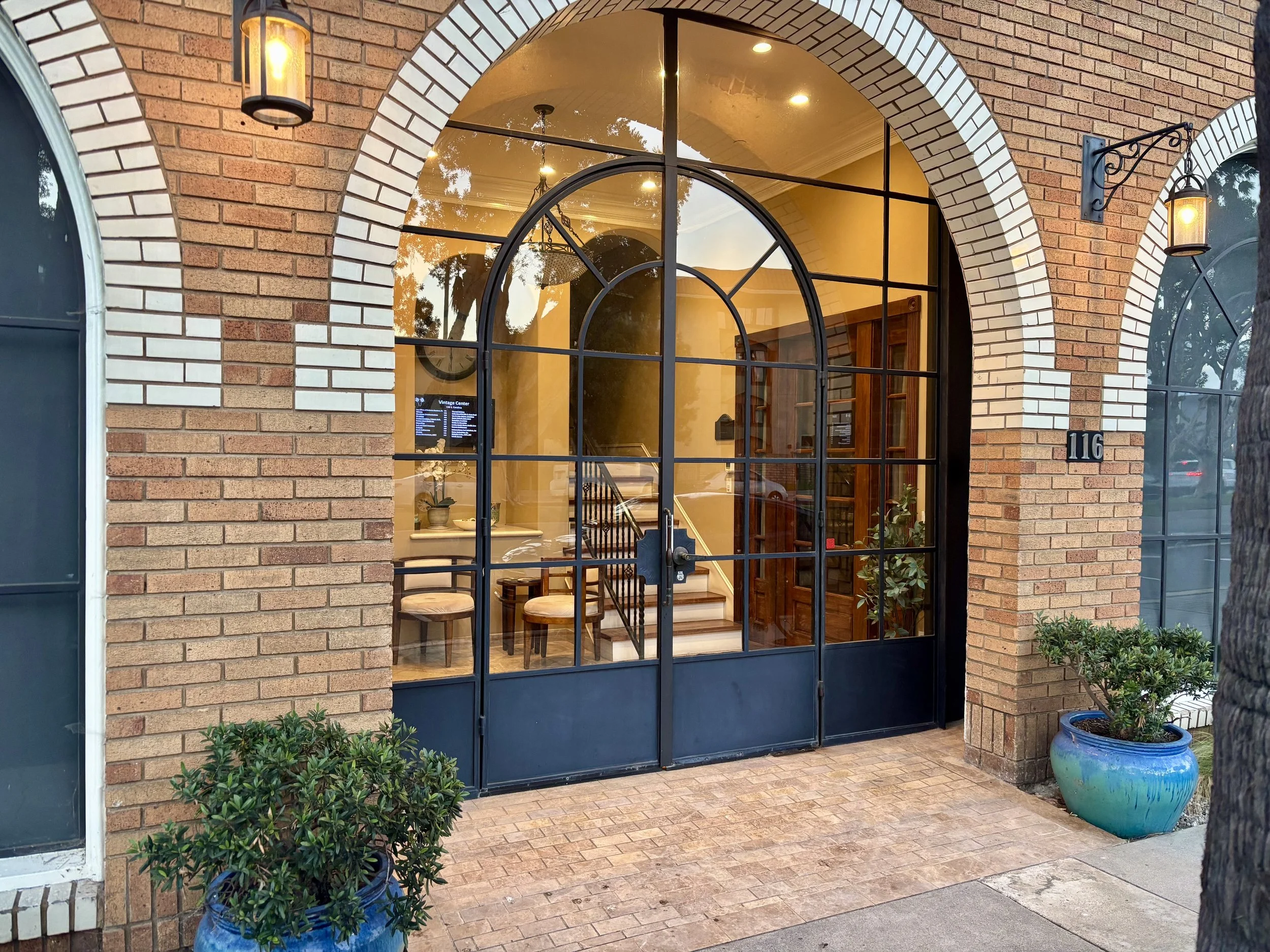 View of a storefront with large arched window and brick exterior. Inside, warm lighting illuminates a cozy interior with wooden shelves and chairs. Potted plants outside the entrance enhance the welcoming atmosphere.