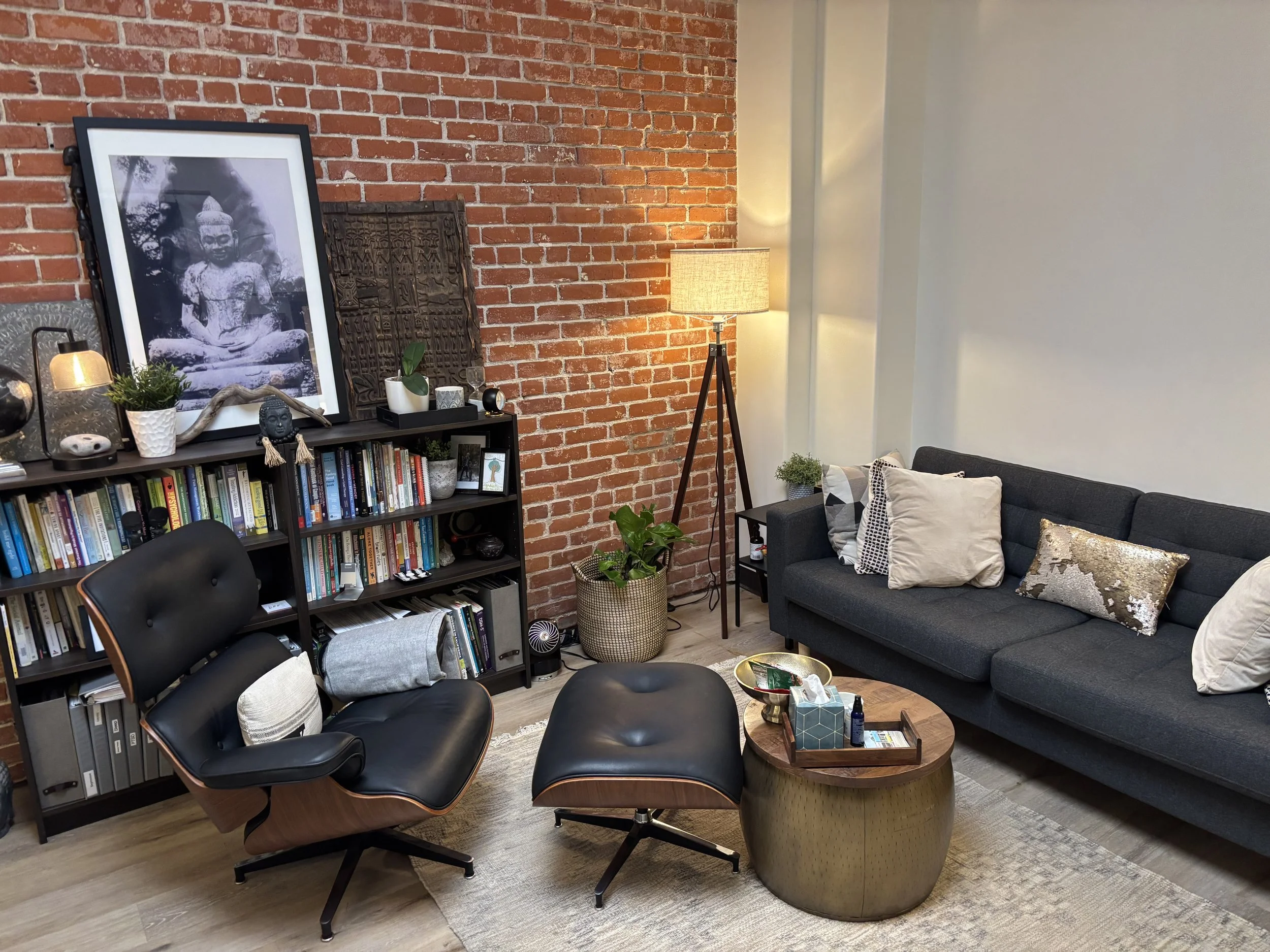 A cozy living room with a brick wall, a gray sofa with various pillows, a wooden coffee table with a tissue box and a tray, a bookshelf filled with books, a tall standing lamp, a potted plant, and a framed black-and-white Buddha statue photograph leaning against the brick wall.
