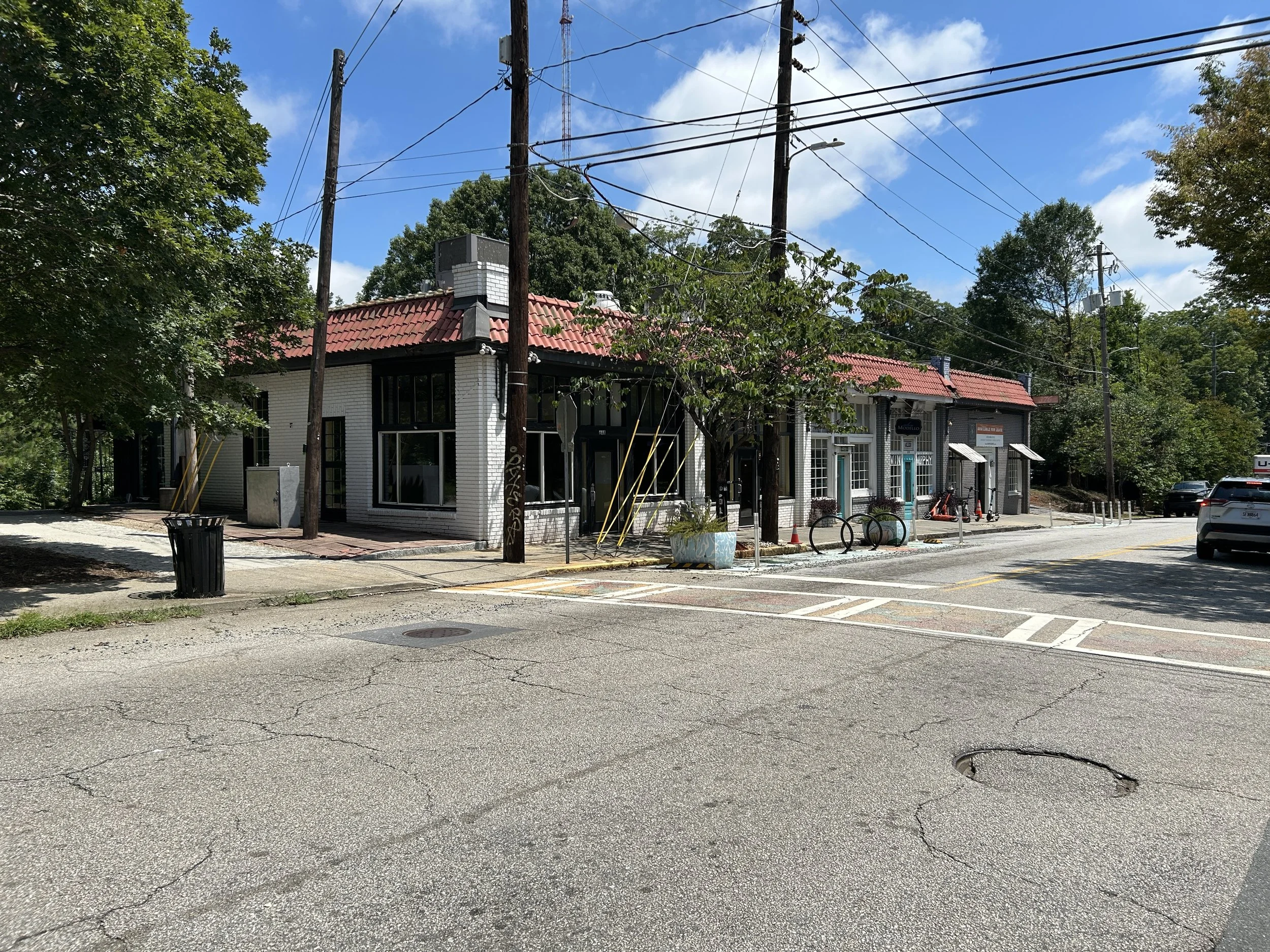 Street view with pavement, a trash can, trees, a white building with large windows and red roof tiles, utility poles and wires, parked cars, and traffic cones, under a partly cloudy blue sky.