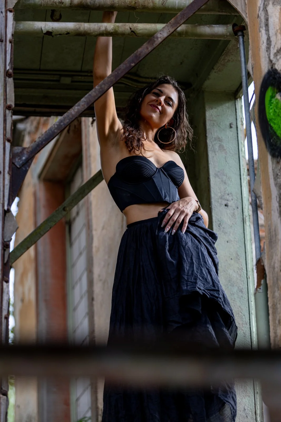 Valerie Sky | A woman with curly hair standing in an abandoned building, wearing a black strapless top and black skirt, posing onfidently..jpg
