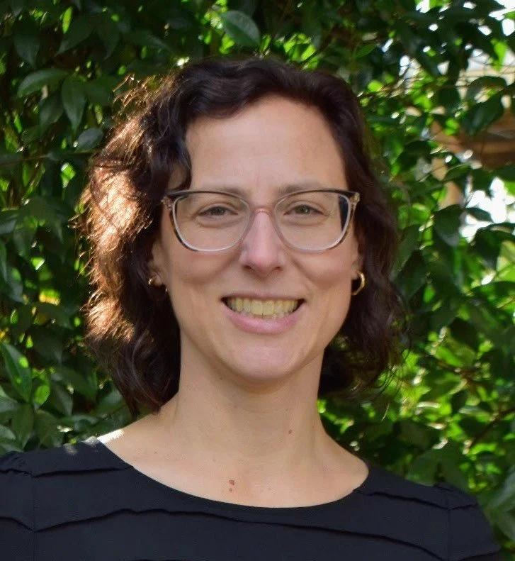 A woman with short dark curly hair and glasses, smiling outdoors in front of green bushes, wearing a black top.