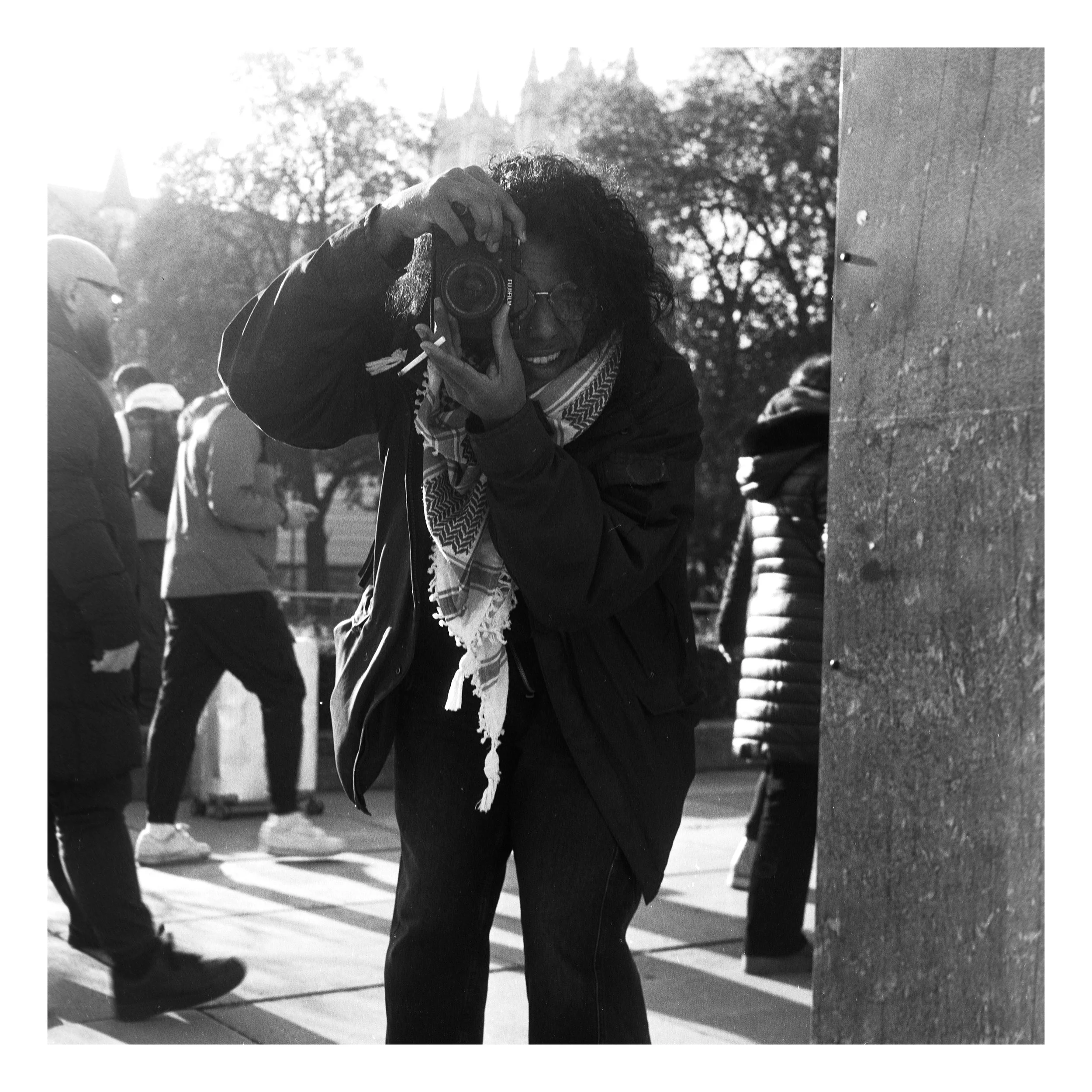 Photo of Tarun holding a camera to their eye and taking a photo. They are wearing a black coat, black trousers, a keffiyeh, and are holding a cigarette in the hand focusing the lens of the camera.