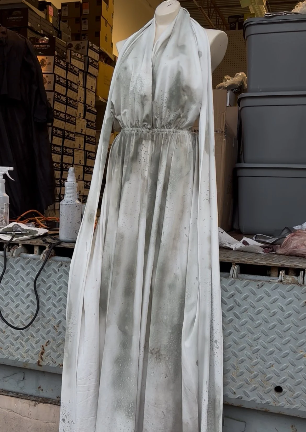 A mannequin dressed in a white, stained, and dirty dress or gown displayed on a platform in what appears to be a storage or warehouse area.