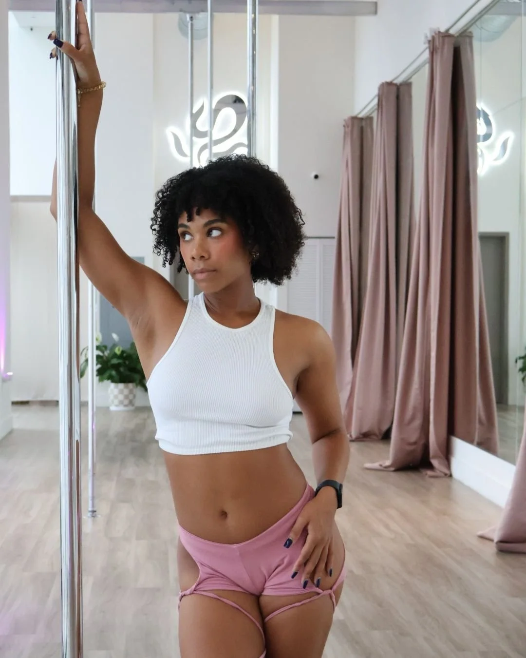 A young woman with curly black hair and medium skin tone posing in a dance studio. She is wearing a white sleeveless crop top and pink shorts with cut-out sides. She is holding a pole with one hand and looking to her left.
