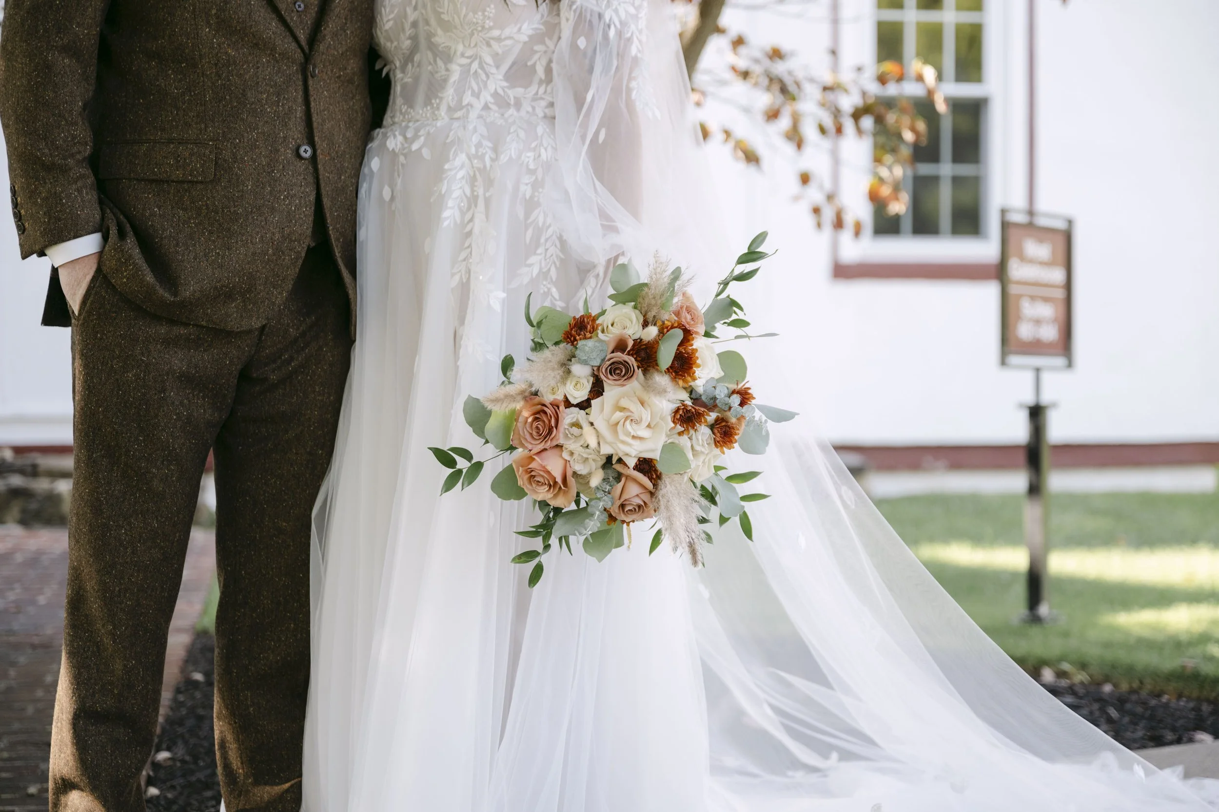 Close-up of a bride holding a bouquet of flowers standing next to a groom, during a wedding ceremony outside near a house with a window and a sign in the background.